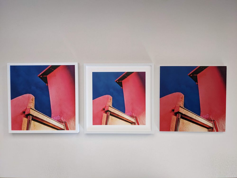 Three framed photos of a modern building with pink walls and a blue sky, arranged side by side on a white wall. The images are nearly identical but vary slightly in color tone and framing.