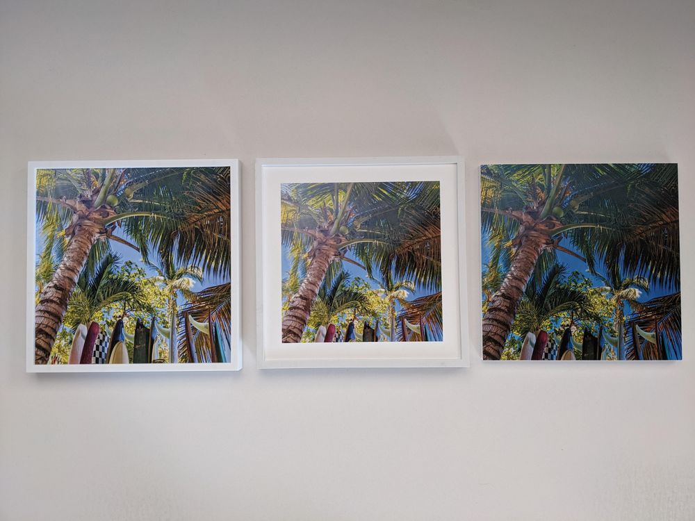 Three framed photos on a wall show tall palm trees and colorful surfboards against a blue sky, viewed from below. The middle frame is slightly tilted.