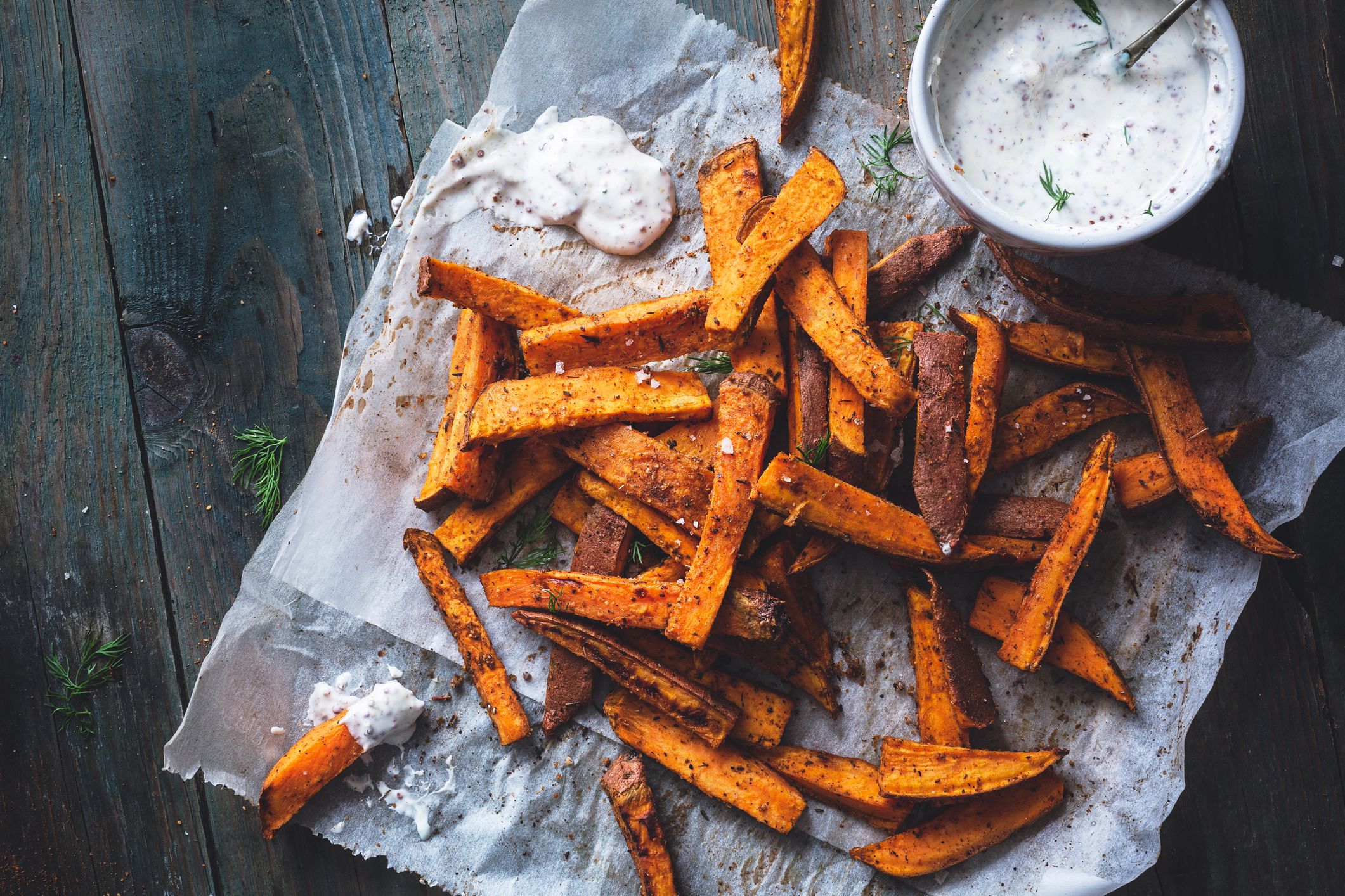Sweet potato fries with cajun spices with yogurt and dill sauce on the side on wrapping paper on a rustic dark grey wooden table