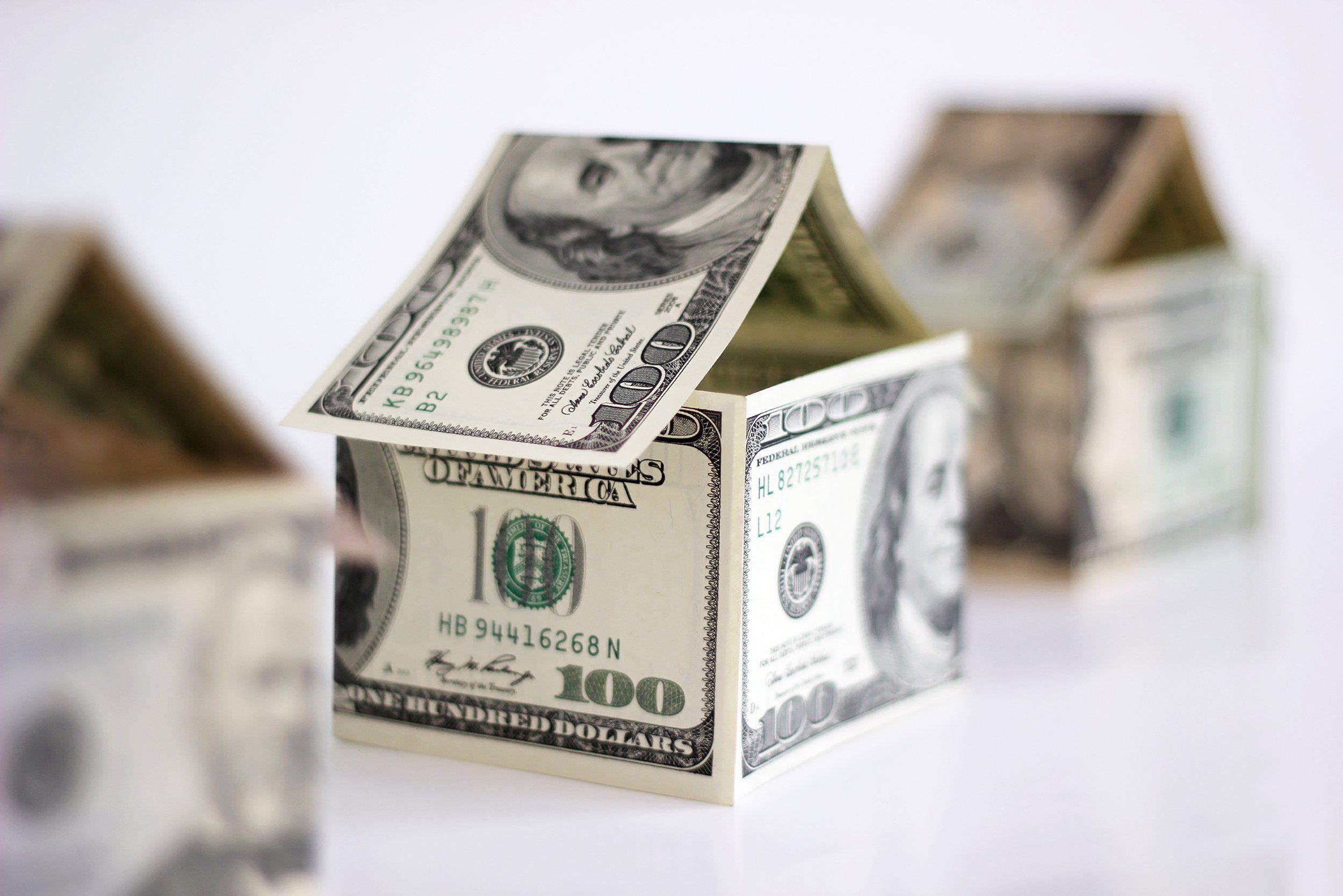 Three houses made out of $100 U.S. Dollar bills, middle one is in selective focus, other two are blurred, on a white table with a white background