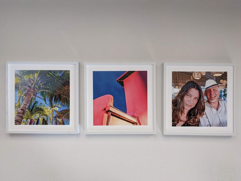 Three framed photos hang on a white wall: a palm tree scene, a red building against a blue sky, and a smiling couple sitting together under lights.