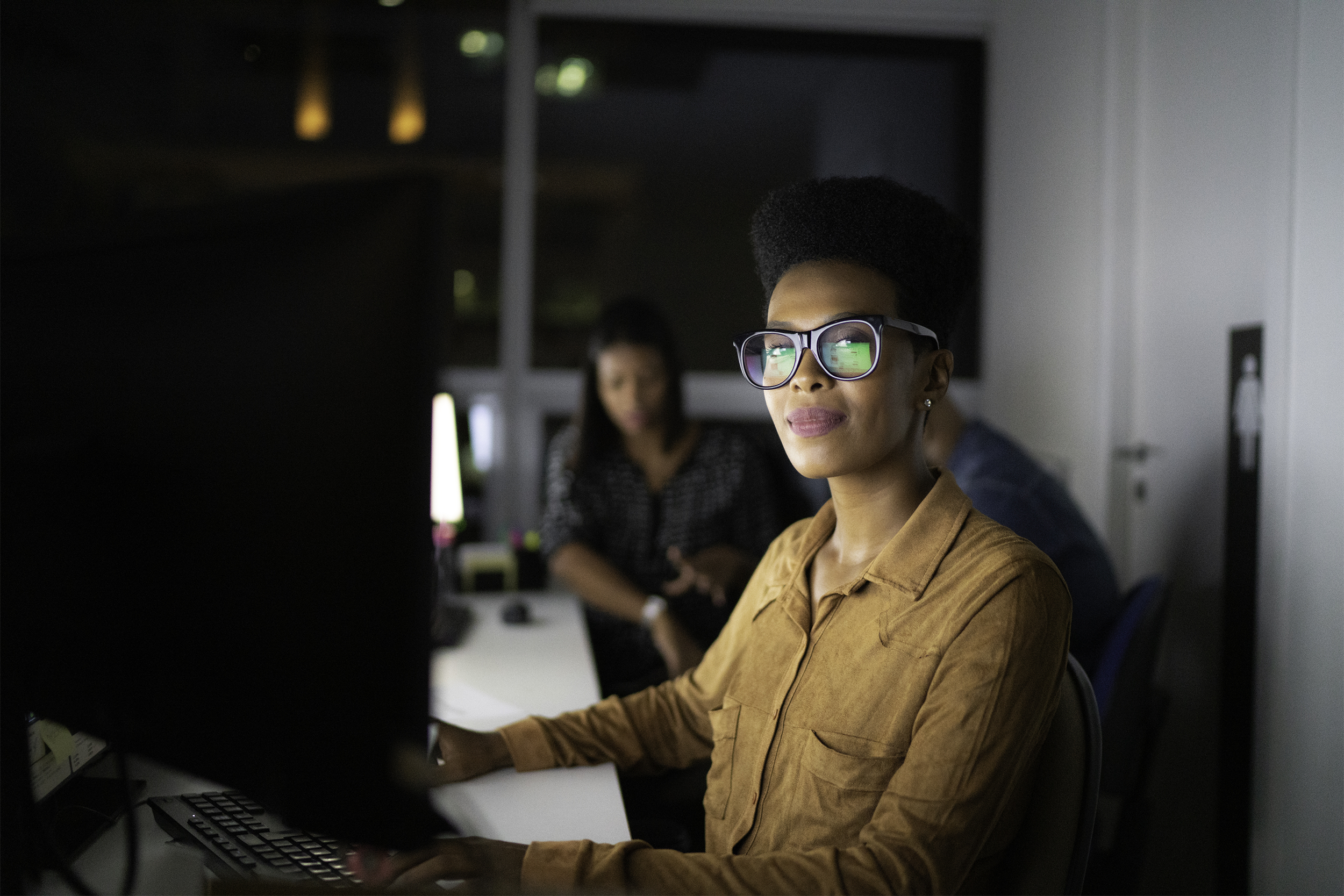 Woman looking at camera while facing her desktop computer at work during the late evening, darkness in the window, a female and male coworker sitting in the background, blurred