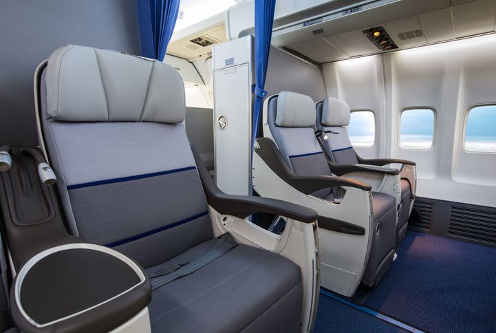 Two spacious, modern airplane seats in a premium cabin, with large armrests and ample legroom, separated by a blue curtain, next to windows showing a clear sky outside.
