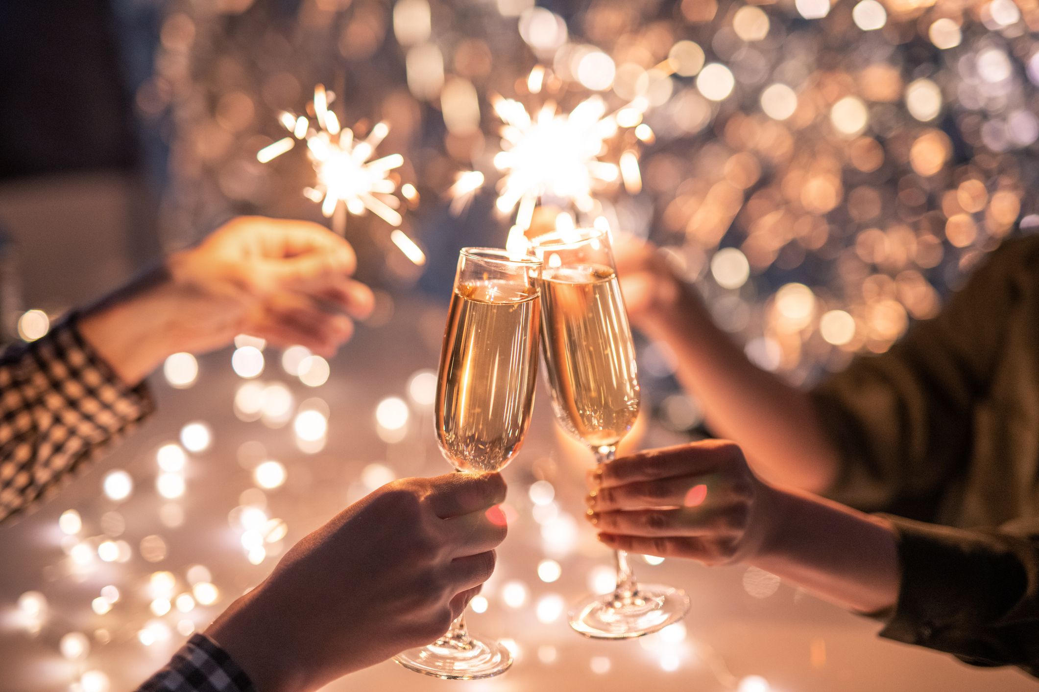 Three people clink champagne glasses together while holding sparklers, with warm, festive lights creating a celebratory atmosphere in the background.