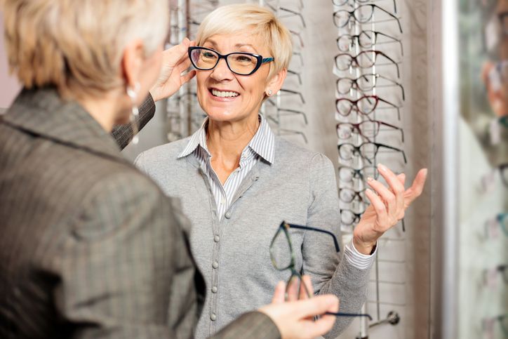 Two older women are in an eyewear store. One woman is smiling while trying on eyeglasses, while the other holds a pair of glasses. Behind them is a display wall with various eyeglass frames.