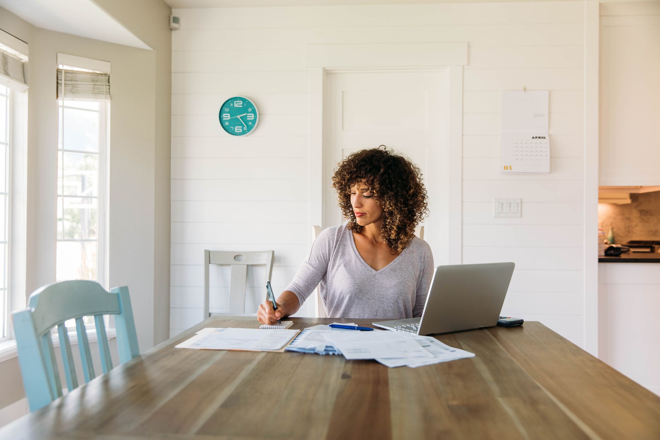 A woman sits at her dining room table with laptop and financial reports doing her monthly budget. She is writing down budgets as she works on her computer to do monthly finances, pay taxes and save money for the future.