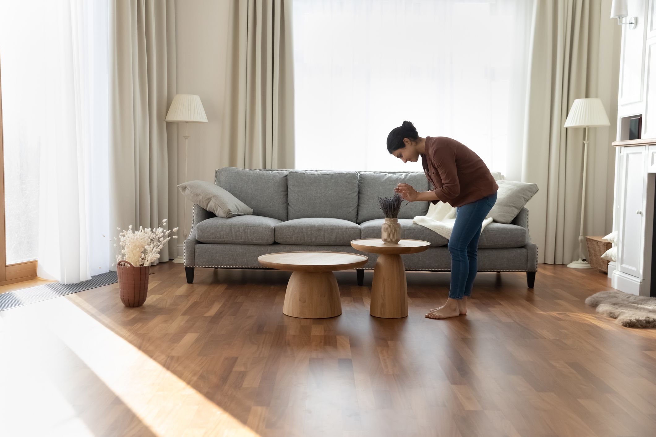 Full length of young Indian ethnicity housewife standing in modern spacious living room create cosiness at home, arranging flowers bunch in vase. Housework routine, interior designer workflow concept