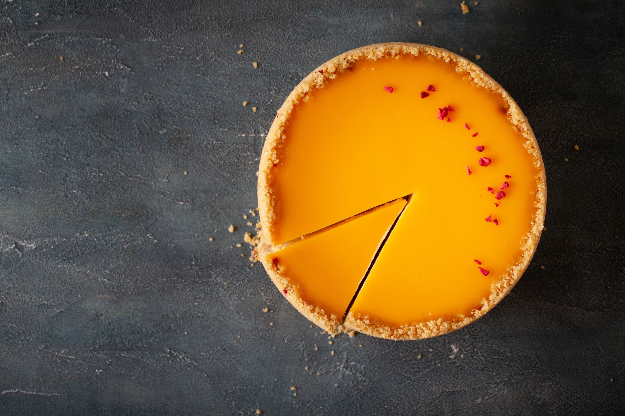 Flat lay of cheesecake with yellow mango flavoured layer decorated with red crumbs of freeze-dried raspberries. Cake cut on the dark marble background