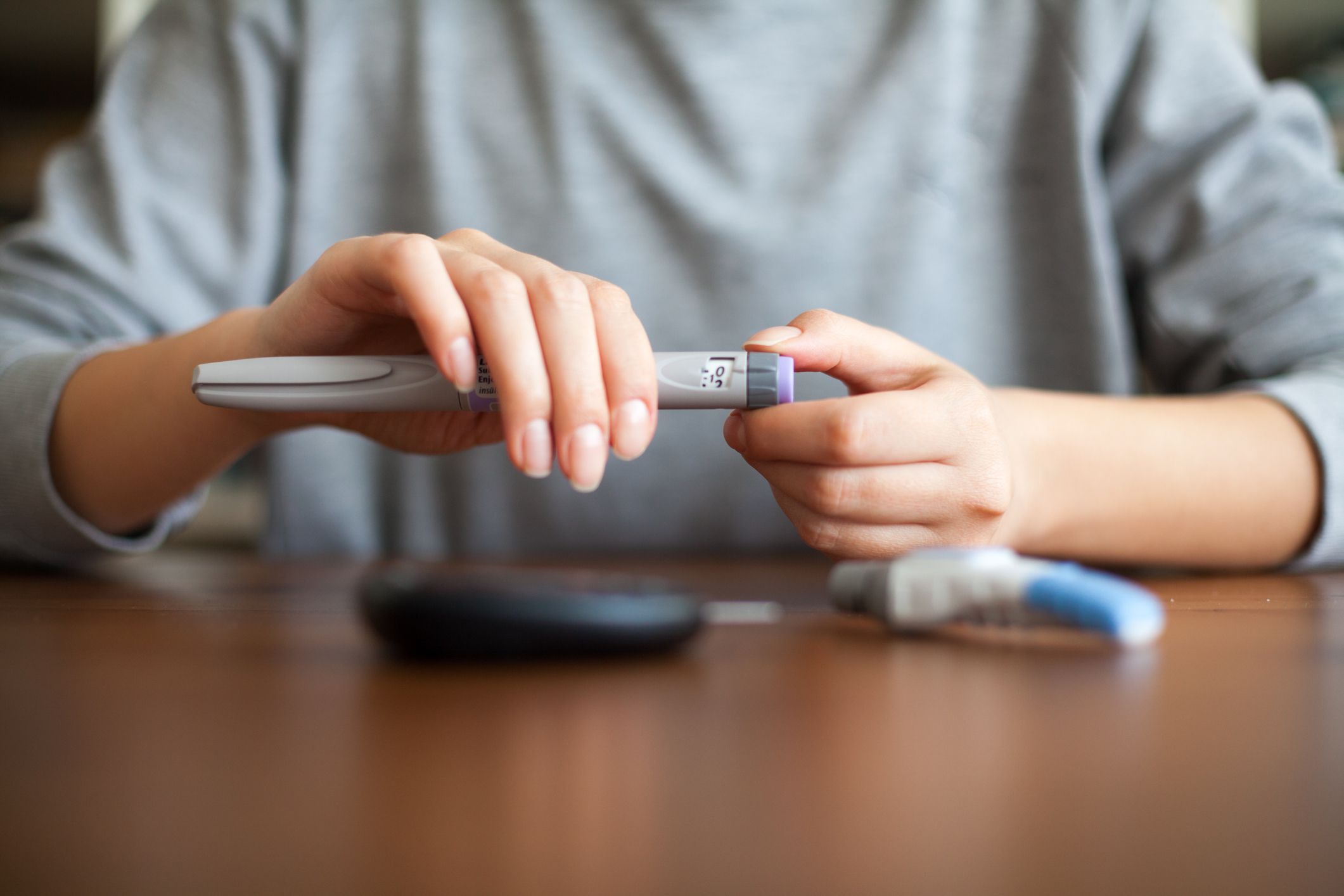 A person sits at a table and prepares an insulin pen by attaching a needle, with a blood glucose meter and supplies nearby on the wooden surface.
