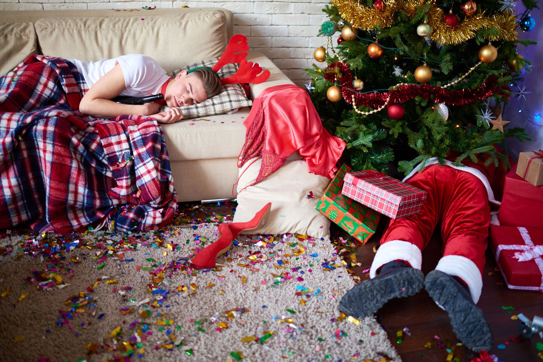 drunken person sleeping with bottle on couch next to christmas tree with fallen santa plush, confetti on carpet