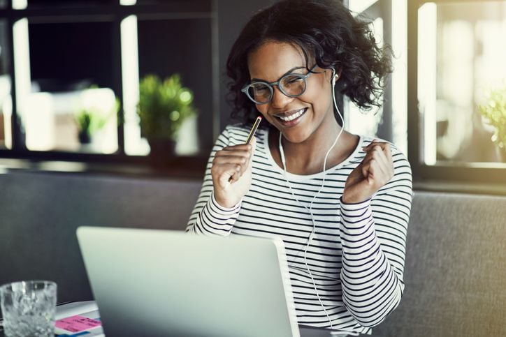 A smiling woman wearing glasses and earphones sits at a table in front of a laptop, raising her fists in excitement. She is dressed in a striped shirt, with plants and sunlight in the background.