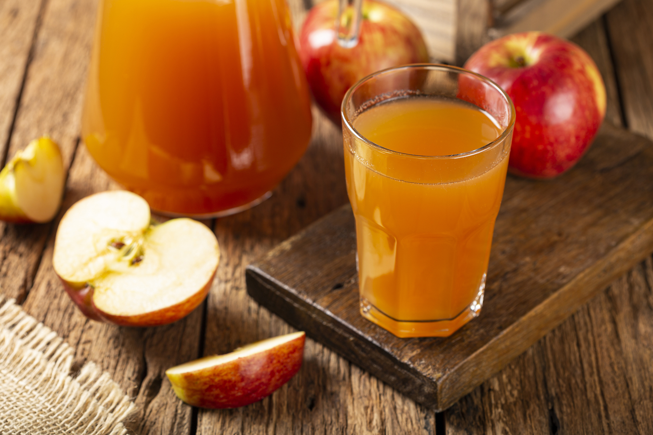 A glass of apple juice, selective focus, on an artisanal wooden board, surrounded by apple slices and a glass jug of apple juice on a rustic wooden table