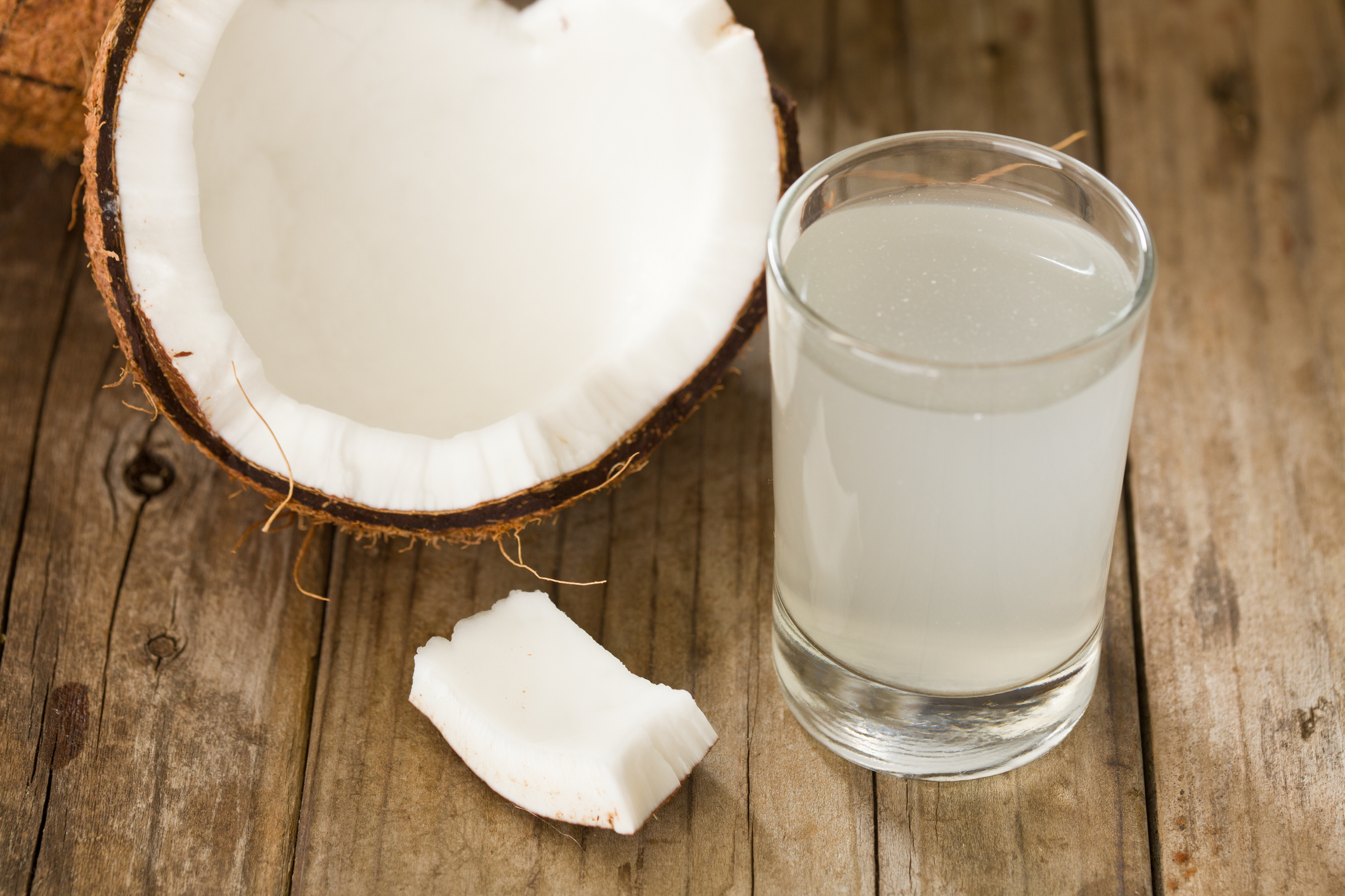 A medium-sized glass of coconut water on the right, a halved coconut, showing white meat on the left, with a piece of coconut meat in the middle on a rustic wooden table