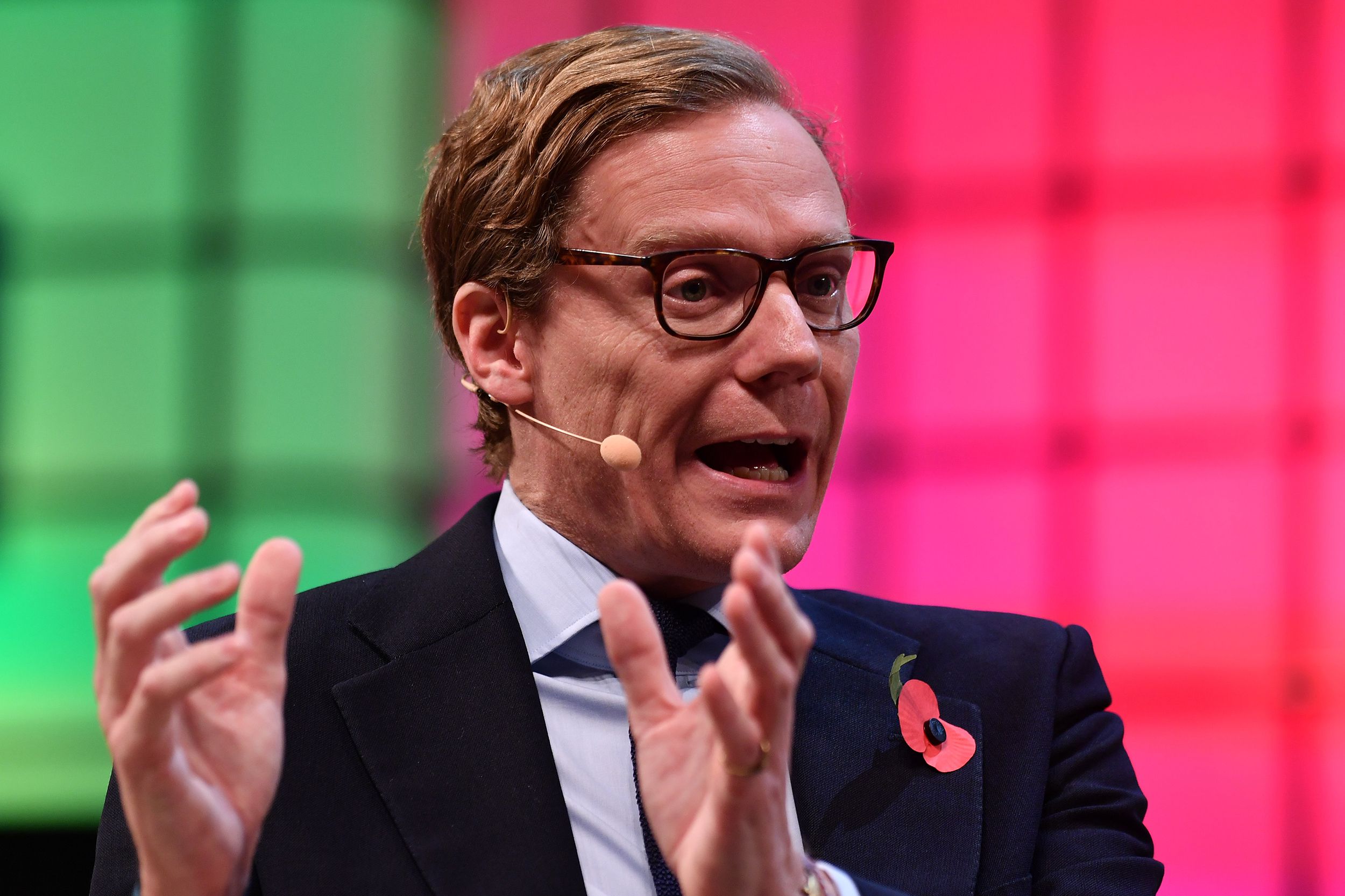 Portrait shot of Alexander Nix giving an interview, while seated, during the 2017 Web Summit at the Parque das Nacoes, Lisbon, Portugal, 2017, a bright lime green and magenta light block screen blurred in the background