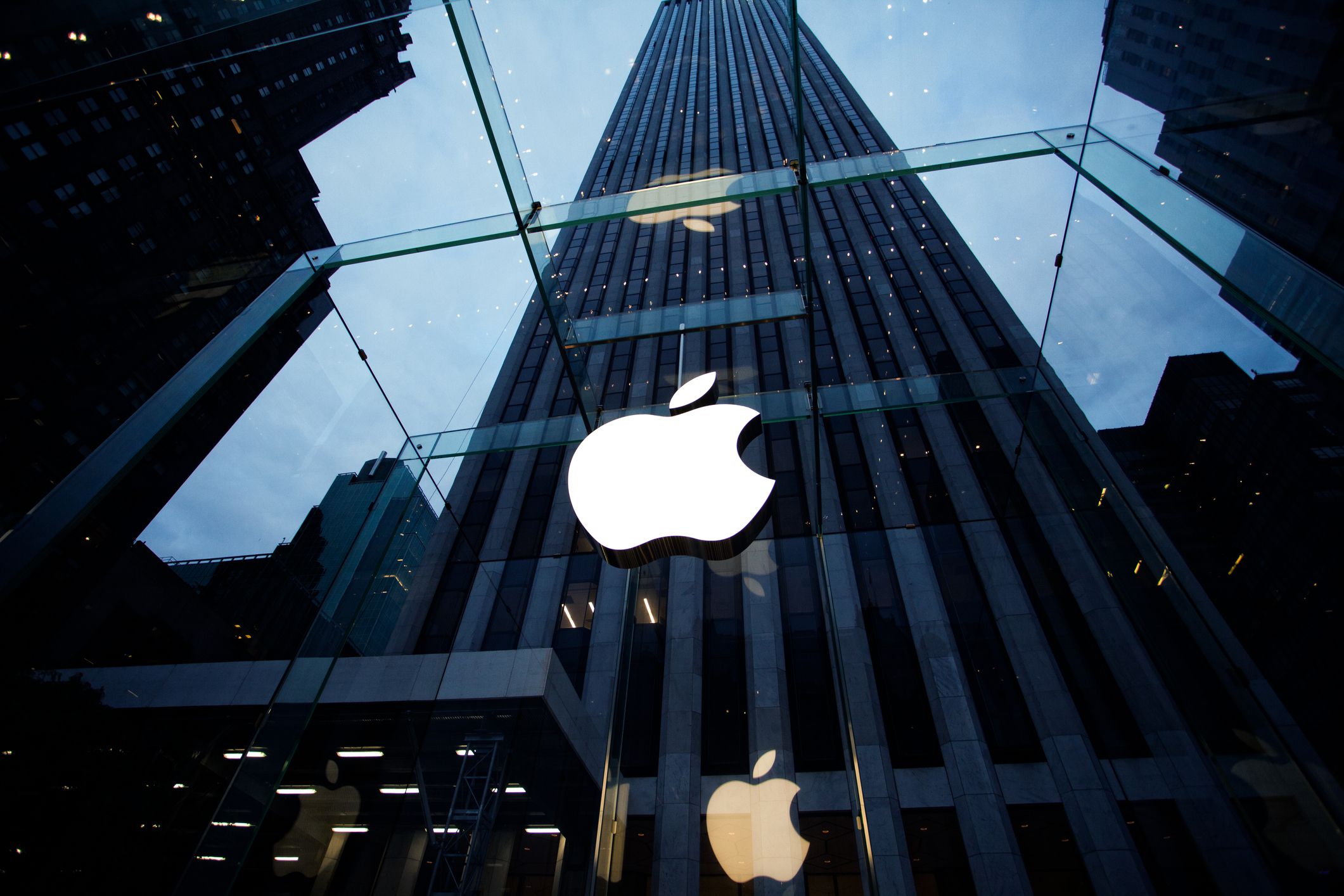 Looking up on the Apple logo in the glass entrance of the Apple store at 5th Avenue near Central Park in New York City with a large skyscraper against a cloudy sky in the background
