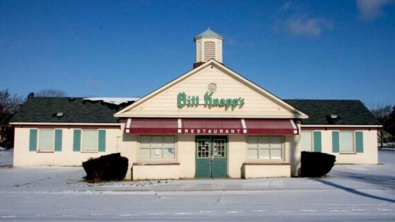 An empty, closed Bill Knapp’s—one of the beloved old restaurant chains—stands with boarded-up windows, a green roof, and central cupola amid snowy ground beneath a clear blue sky.
