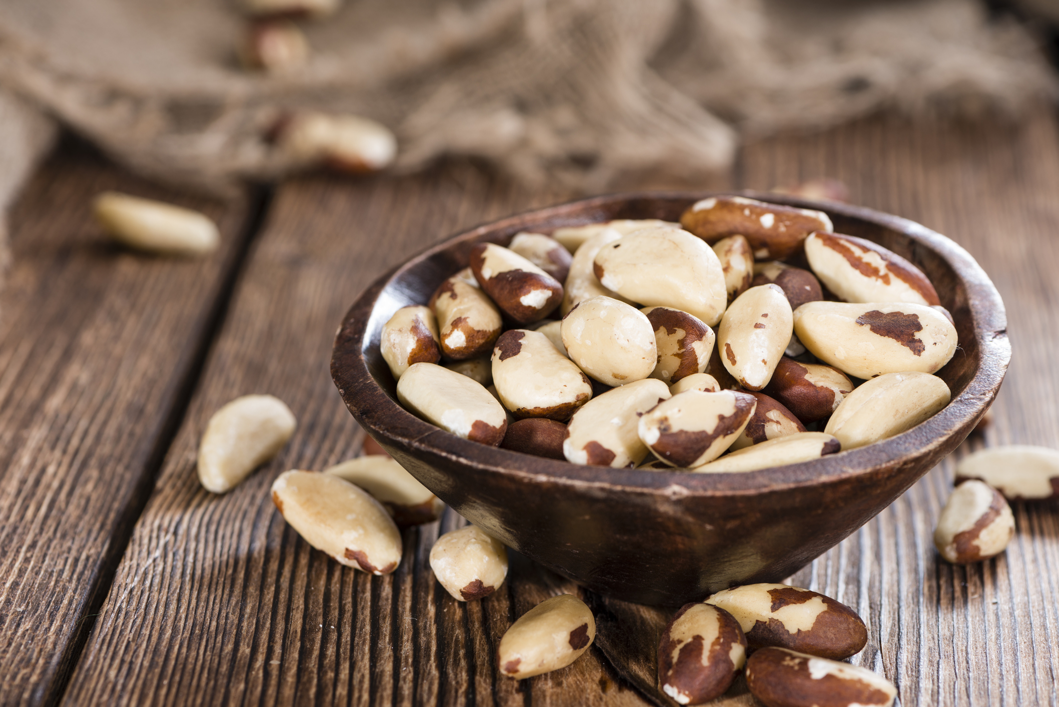 Brazil nuts in a small rustic ceramic brown bowl, surrounded by more Brazil nuts, selective focus, on a wooden table with a burlap bag in the blurred in the background
