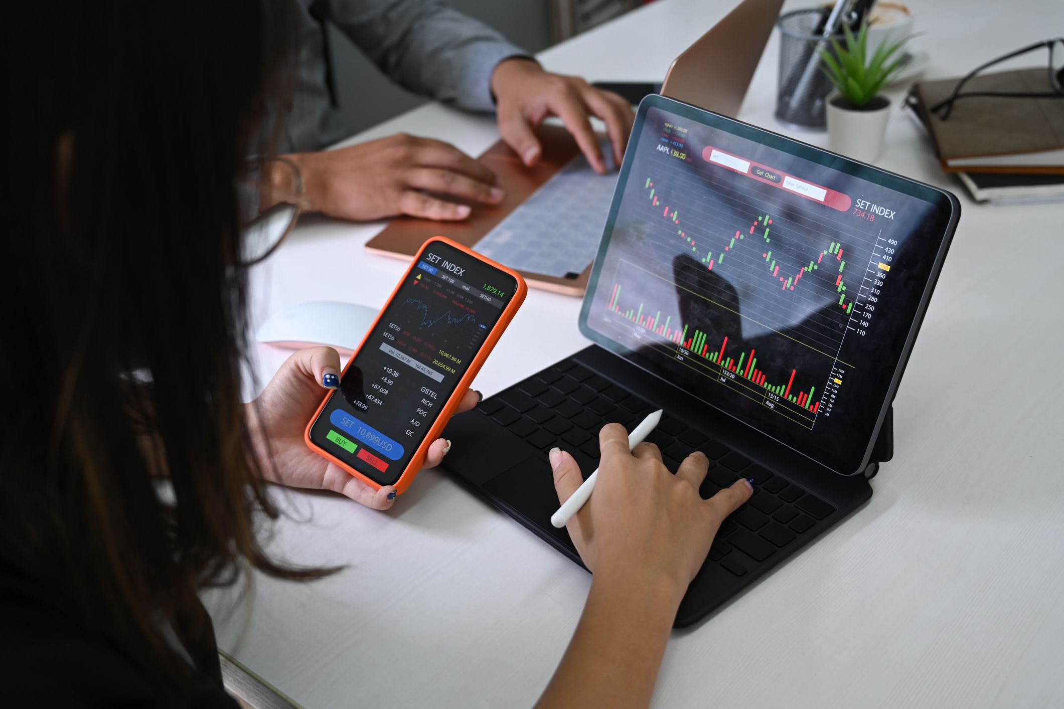 Businesswoman analyzing graphs on computer tablet and smartphone, sitting at a table with male colleague who is on laptop, she is also holding a pen