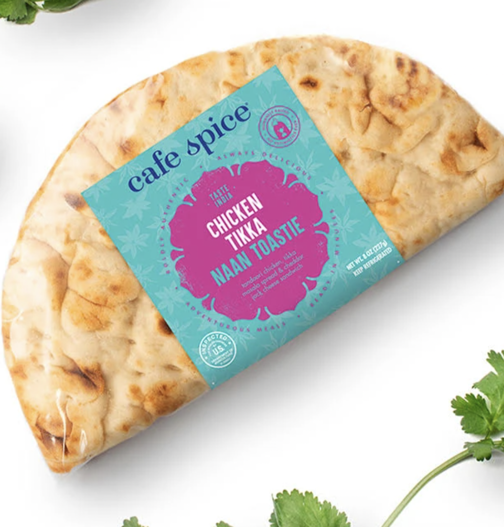 A packaged Cafe Spice Chicken Tikka Naan Toastie sits on a white surface, with fresh cilantro leaves scattered around it. The packaging is turquoise with pink accents and product information.
