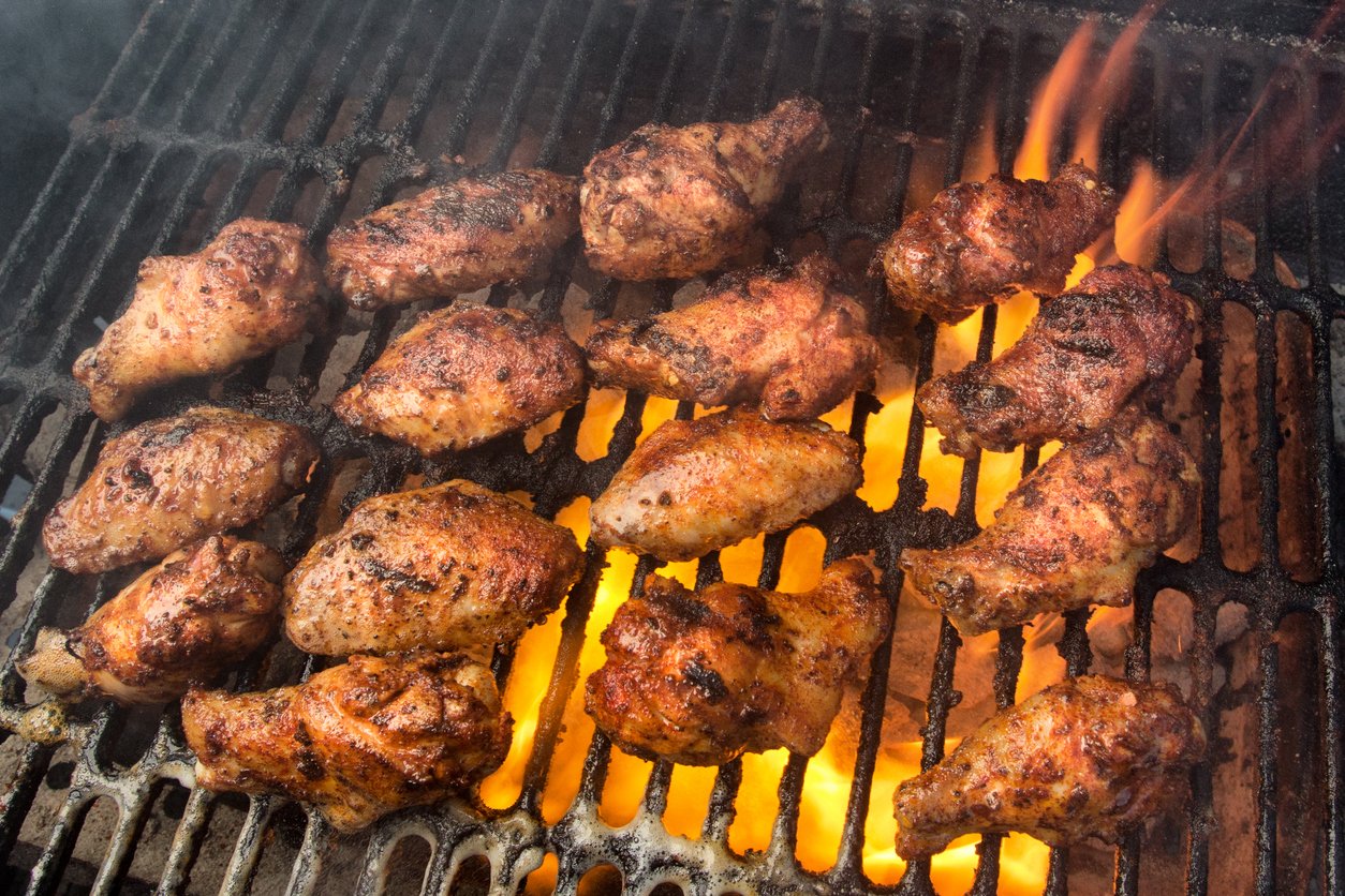Chicken wings being grilled over an open flame on a barbecue, with smoke rising and flames visible between the metal grates. The wings are seasoned and appear crispy and charred.
