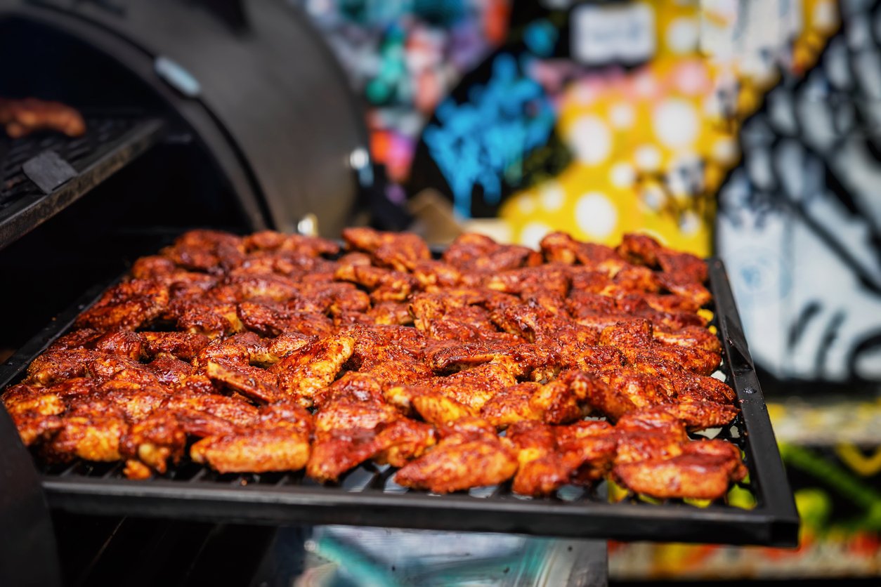 A tray filled with seasoned chicken wings is being placed into a smoker grill, with colorful graffiti art visible in the blurred background.