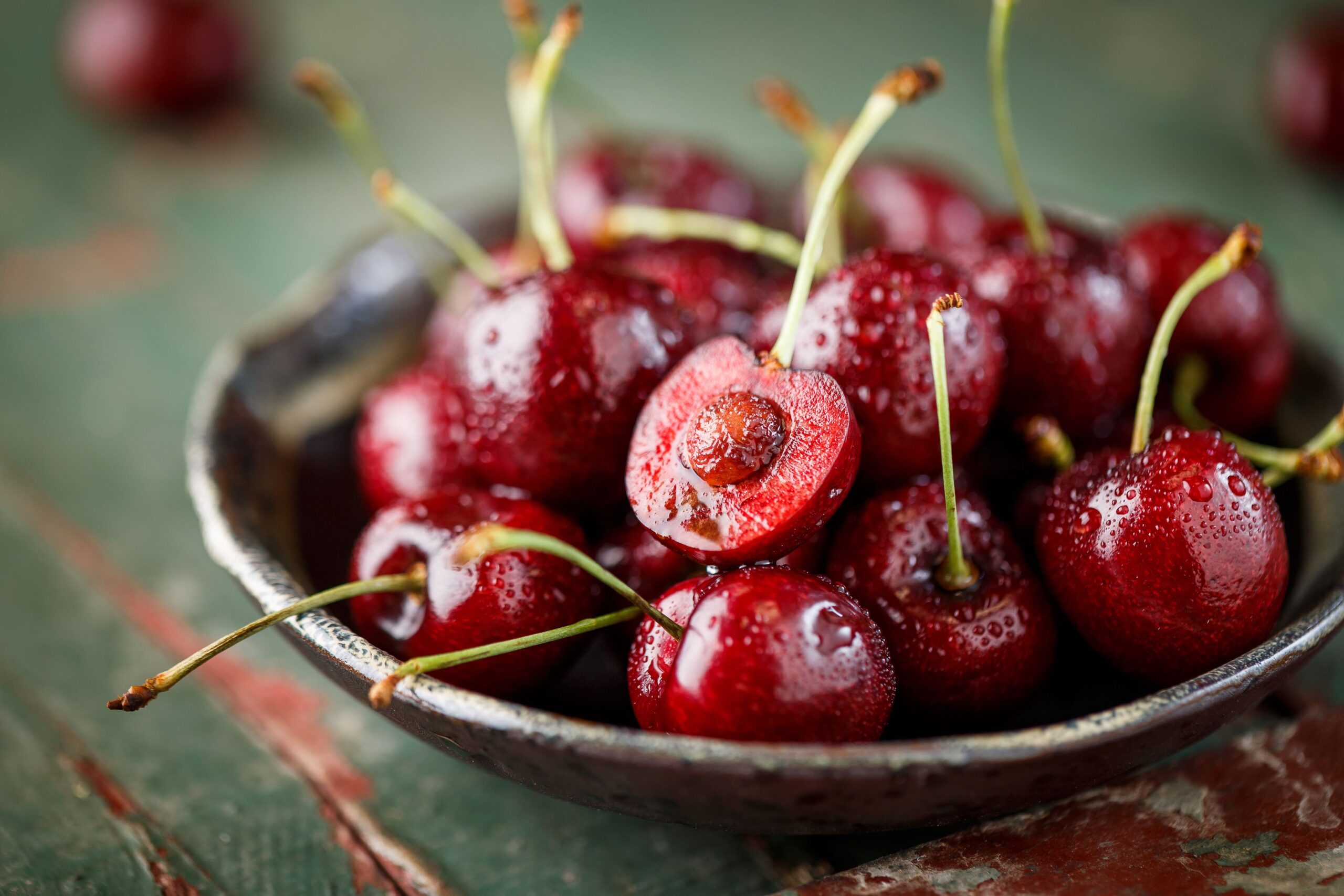 Closeup of cherries in a ceramic bowl, one halved cherry on top, selective focus, still with stems going in all directions on a red-lined green table mat, blurred background