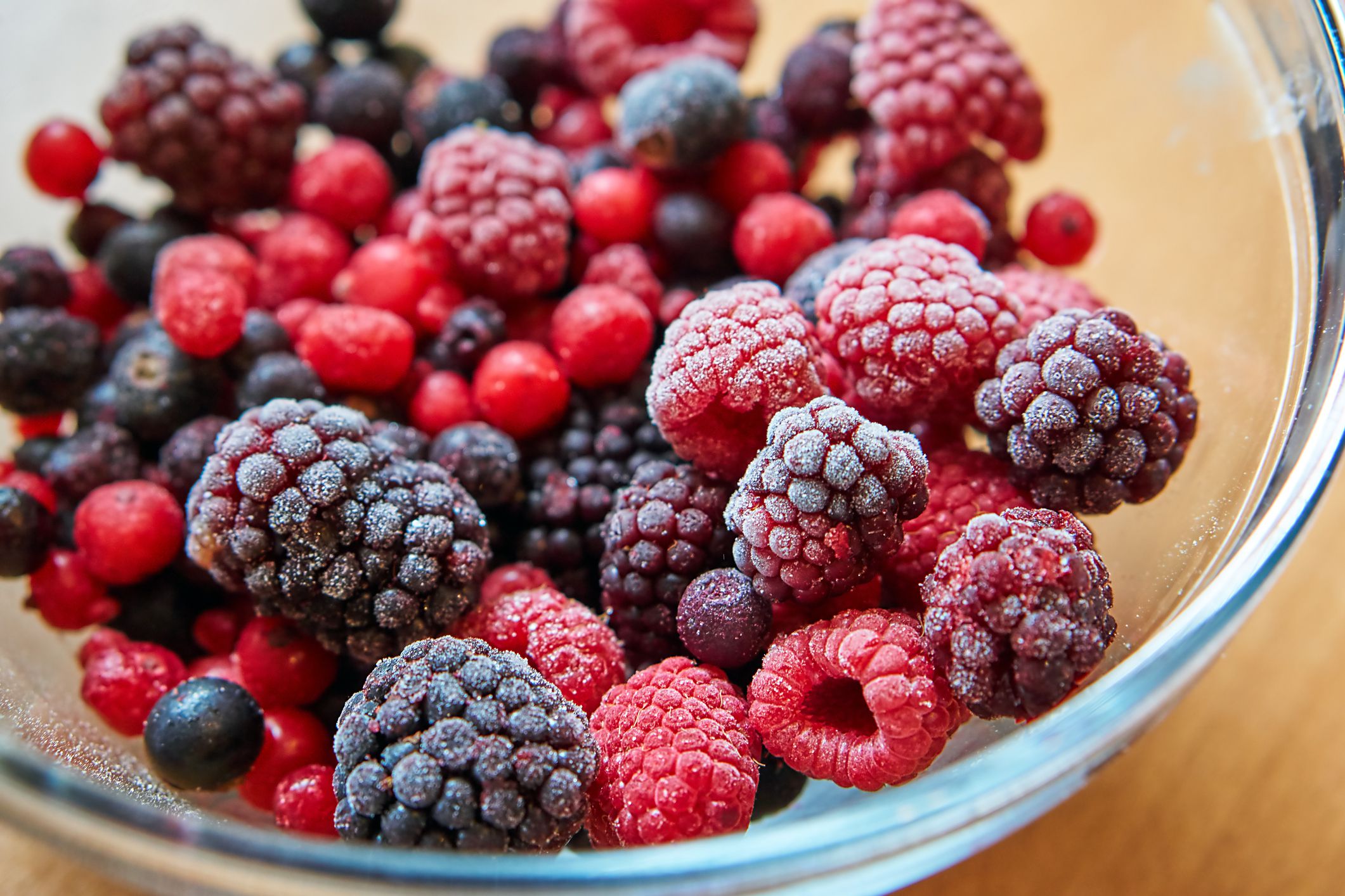 Closeup of raspberries, blackberries, blueberries, and red berries, slightly frozen, in a glass bowl on a wooden table