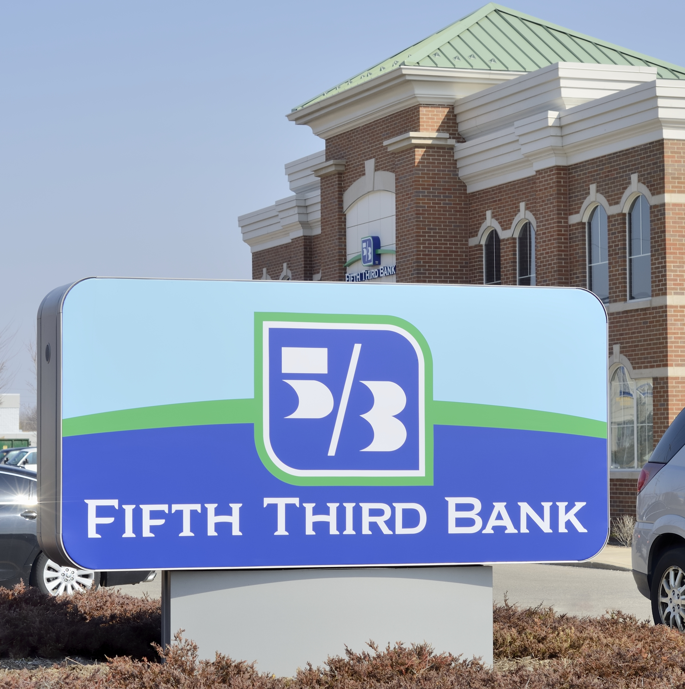 Closeup of sign in the foreground of Fifth Third Bank branch in Rochester Hills, Michigan with building in the background with parking lot and against a blue sky