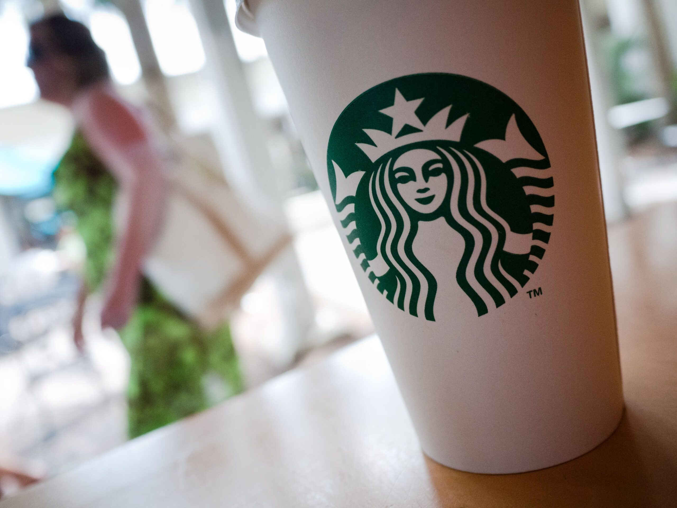 Closeup of Starbucks grande hot beverage cup diagonal on the right on a table in Naples Florida with a woman wearing a green dress and windows blurred in the background