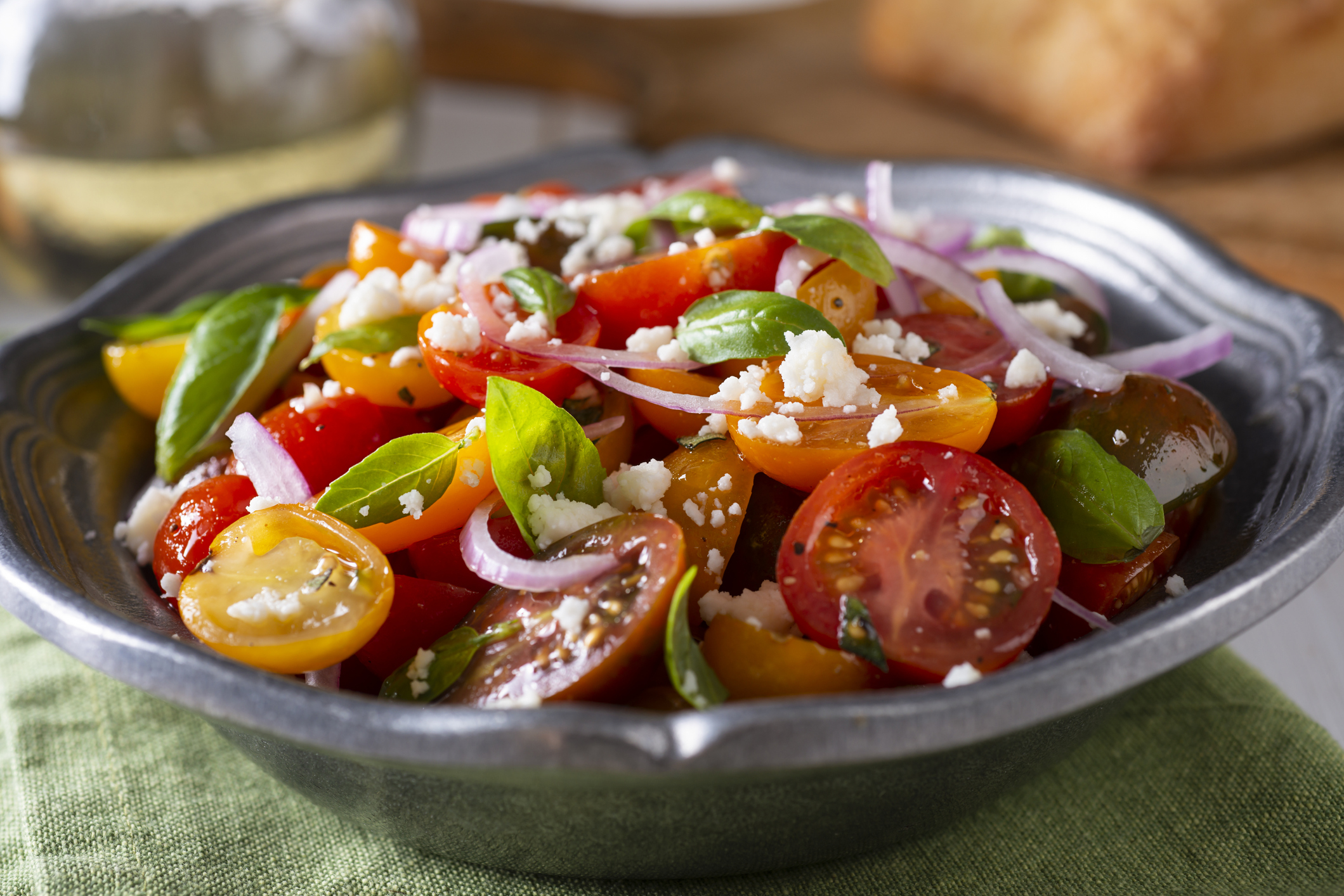 Closeup of tomato salad with onions, herbs, and feta cheese, in a blue ceramic bowl, selective focus, on a sage green napkin, with a blurred background of items on a table