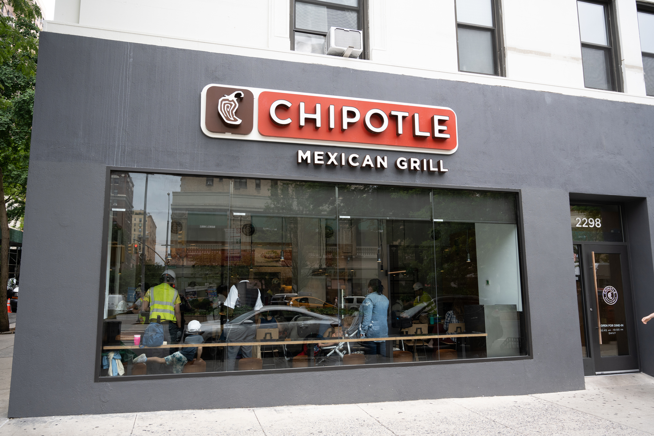 Closeup of a Chipotle Mexican Grill restaurant in New York City, front window and door with front sidewalk and street with trees on the left