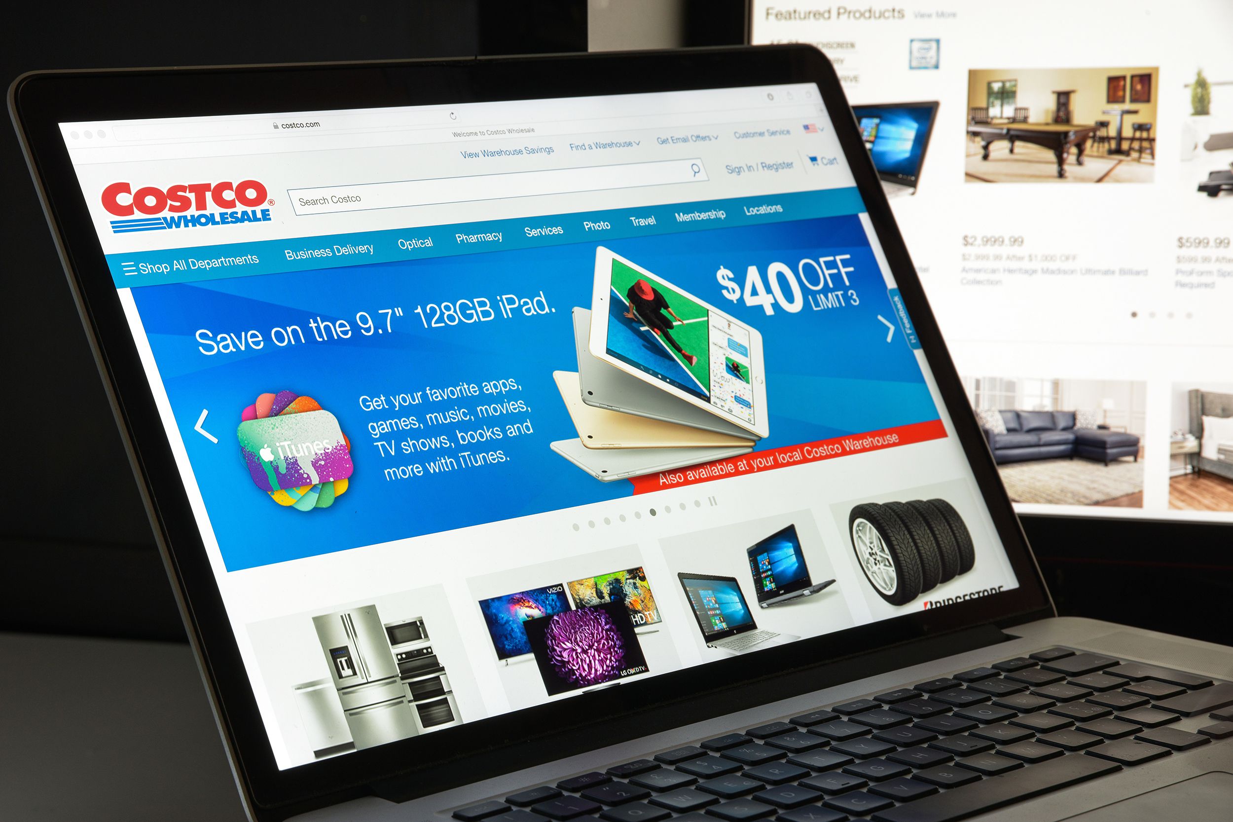 Costco.com website homepage on Safari browser on a MacBook laptop, keyboard shaded, with a desktop computer also showing deals on the Costco website