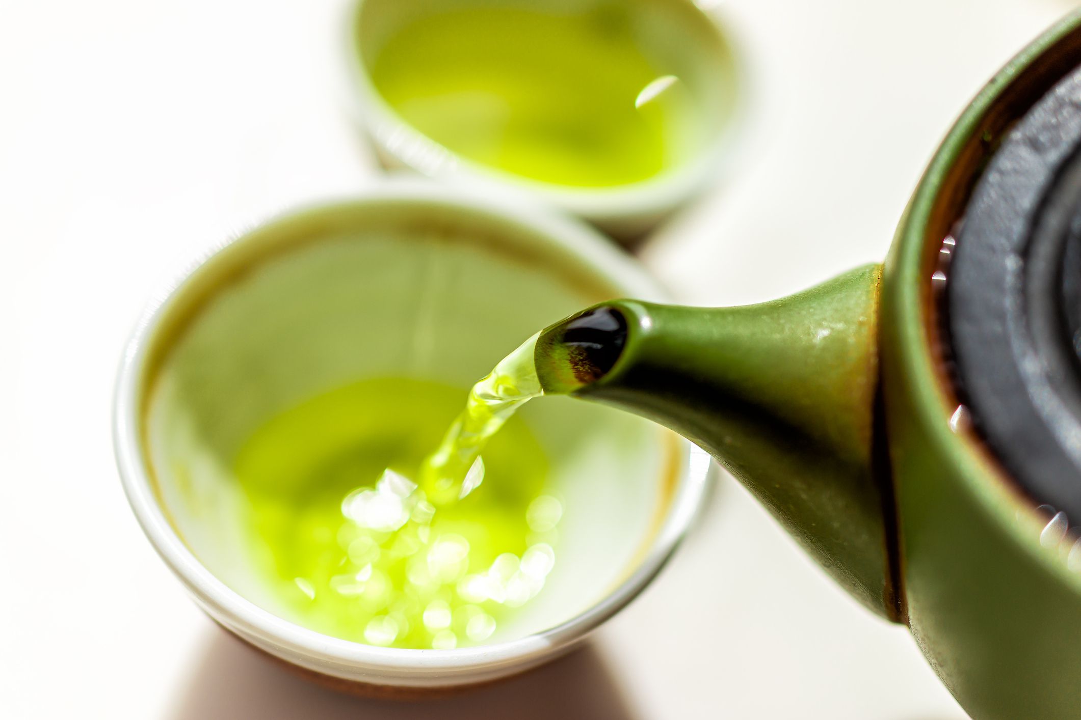 Dark green tea kettle pouring green tea, selective focus, into beige lined white ceramic asian tea cups, blurred, on a white background