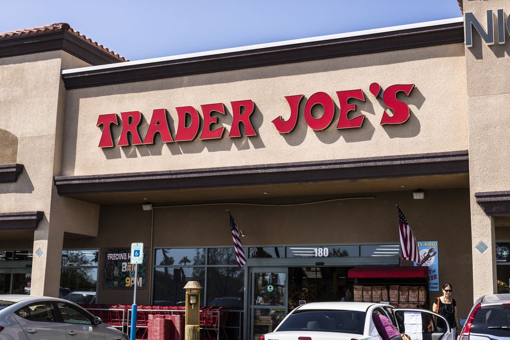 The exterior of a Trader Joe’s grocery store with bold red signage, American flags, and shopping carts visible near the entrance; several cars and a person are in the parking lot.