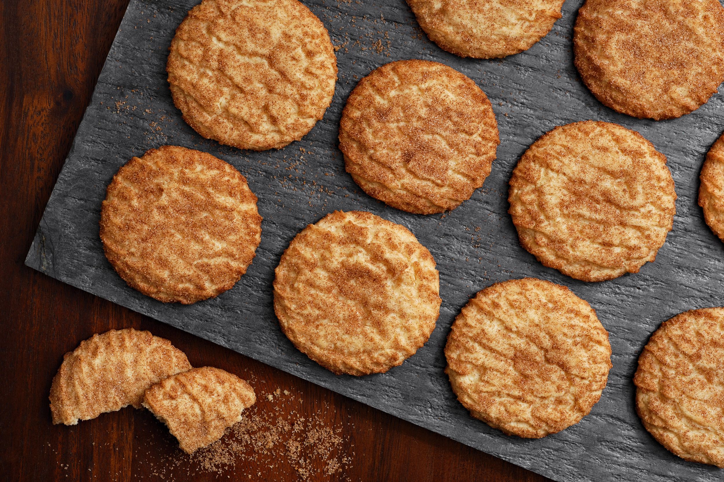 Several diabetic friendly snickerdoodle cookies on a black slate board with one cookie broken into two pieces and snickerdoodle sugar on a wooden table