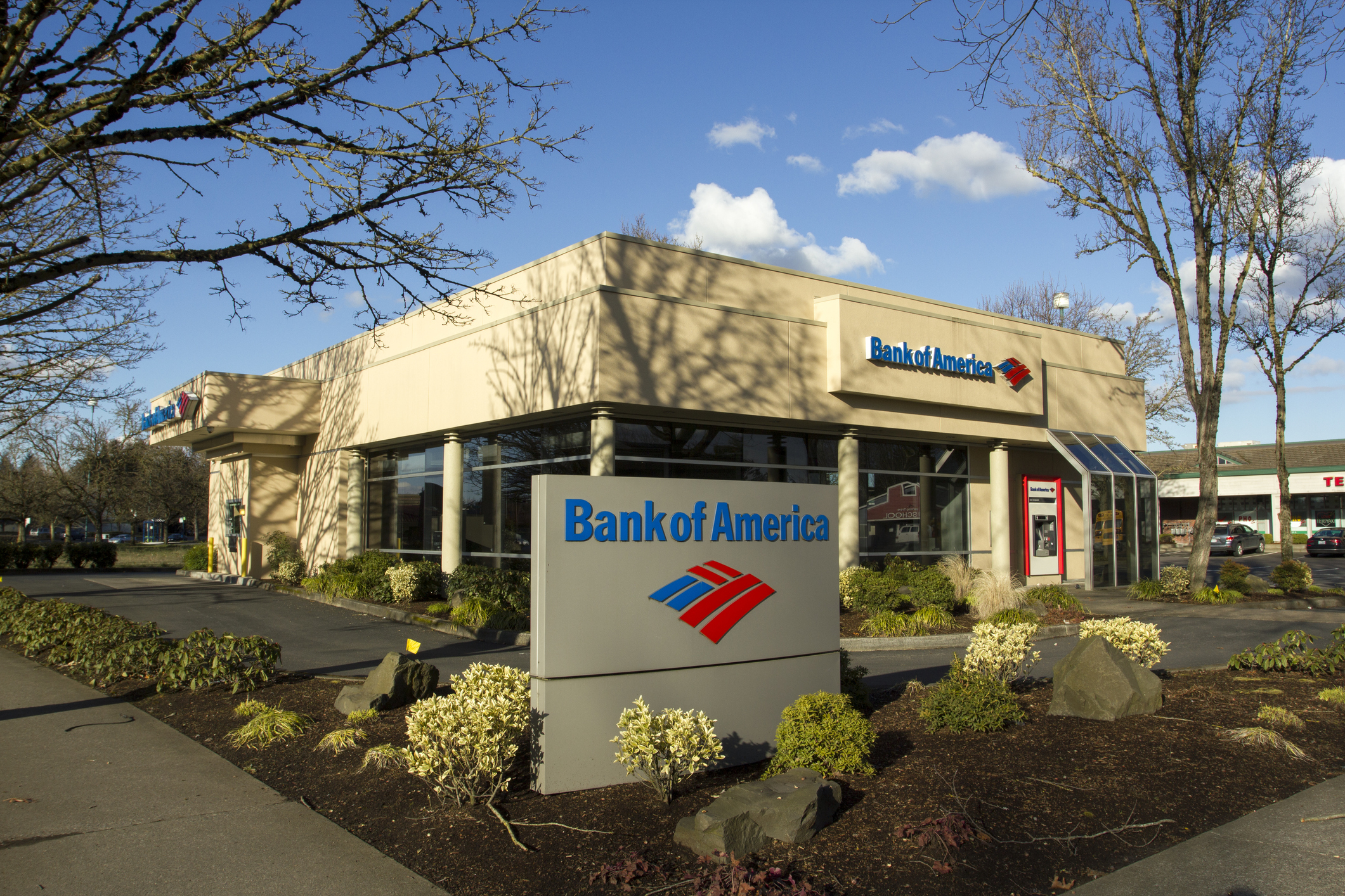 Exterior of a Bank of America branch in Wilsonville, Oregon with sign in the foreground and building in the background, framed by trees, with shopping center in the distant background