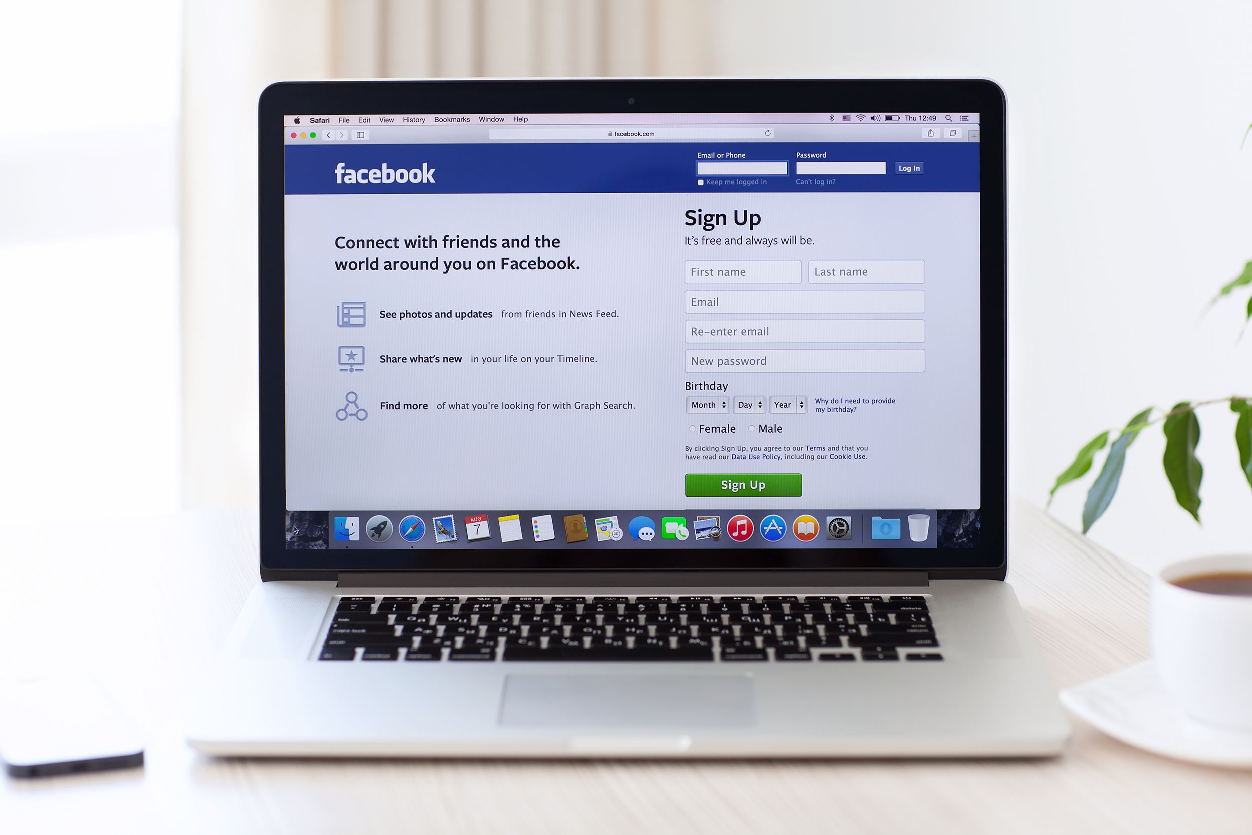 Focus on Facebook homepage on Safari browser on a silver Apple MacBook Pro, in the middle of image, on a light brown desk, surrounded by a blurred background of wall and light coming through window