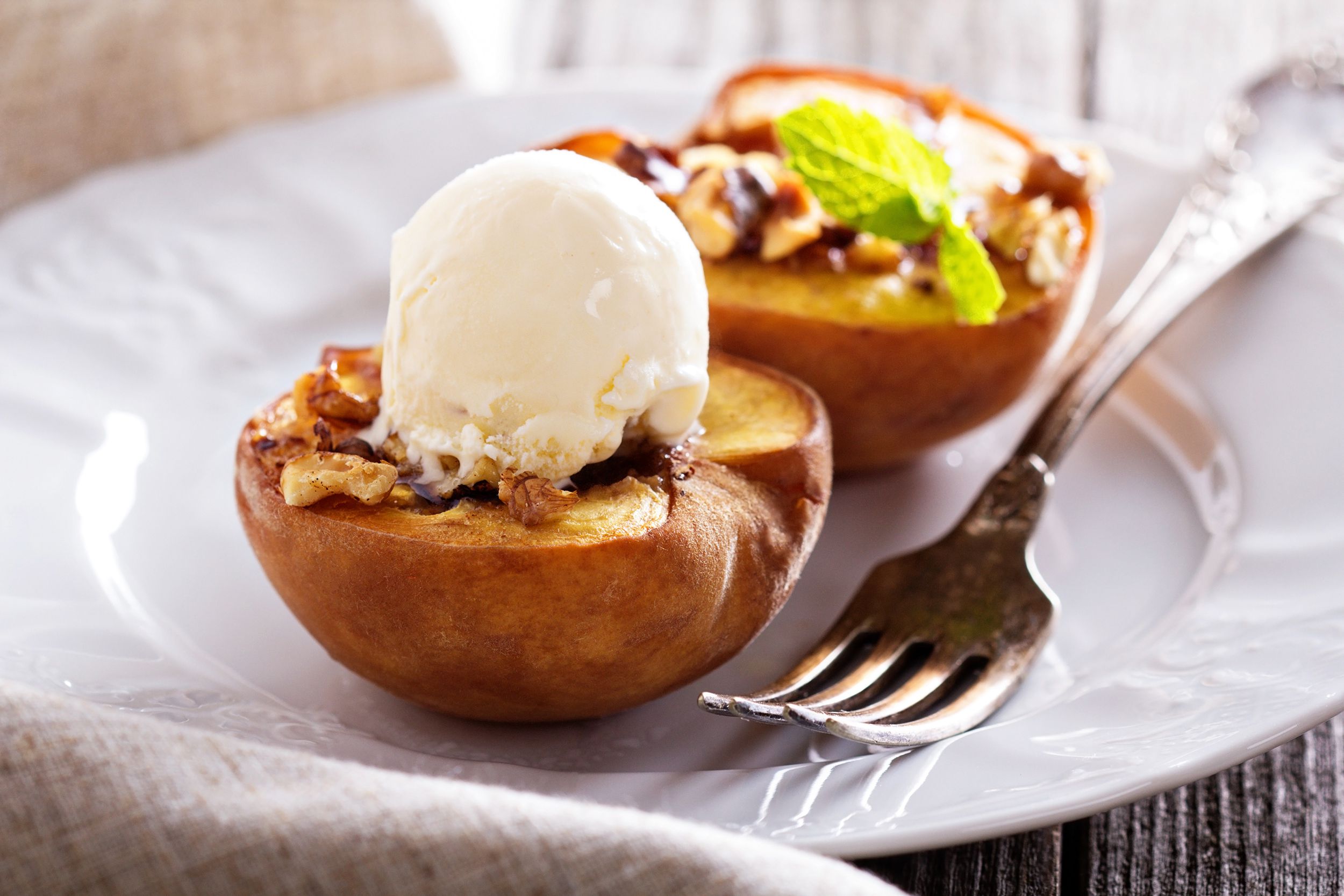Two grilled peaches on a white plate with a fork with a blurred background of napkin and wooden table