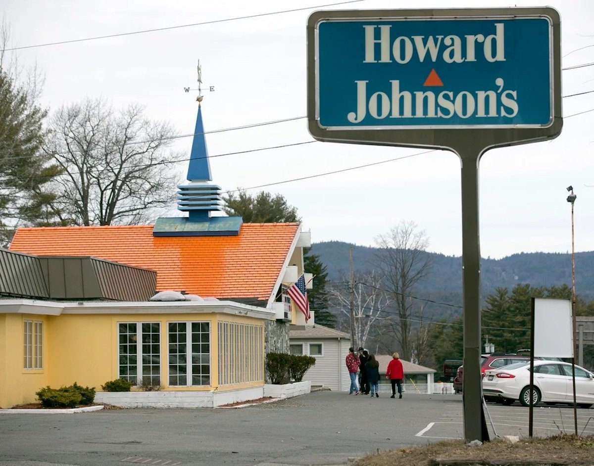 A classic Howard Johnson’s, one of the beloved old restaurant chains, features an orange roof, a blue spire, and a large sign. Several people walk near parked cars, with trees and hills in the background on a cloudy day.