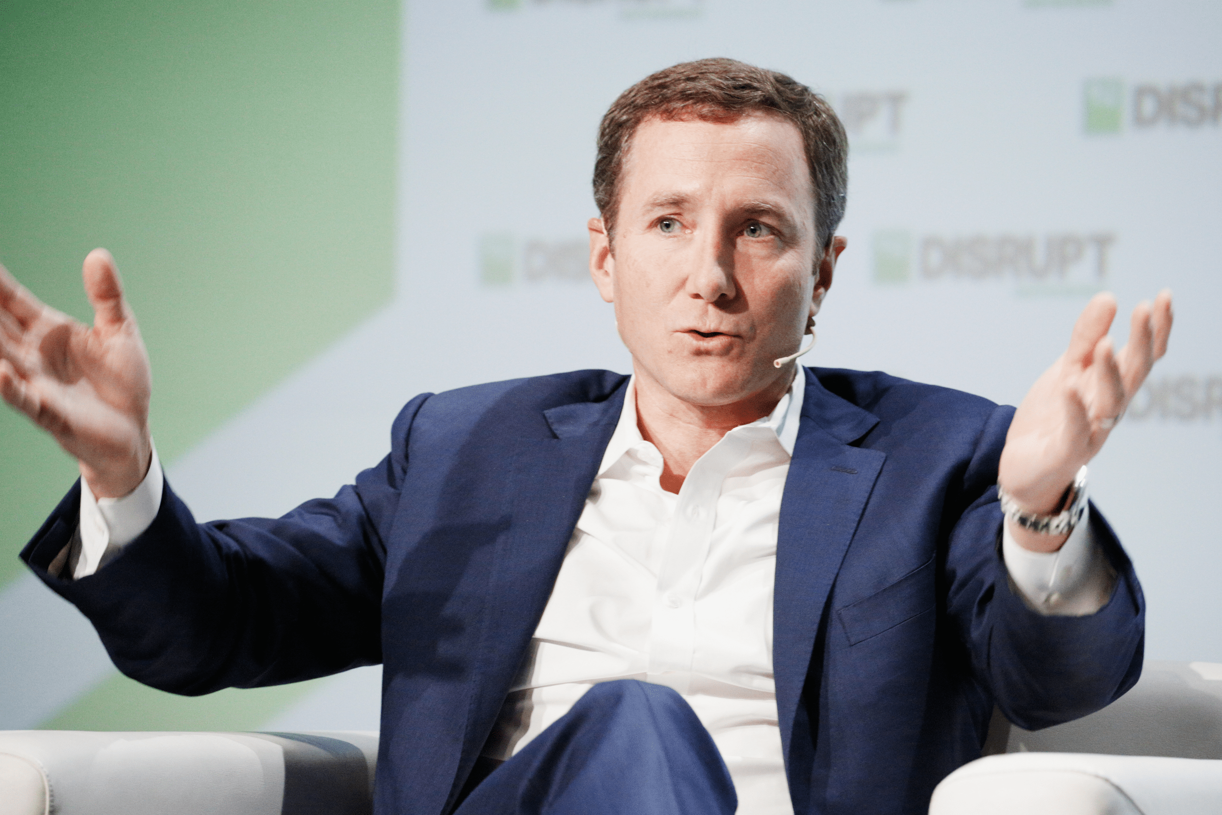Portrait shot of John Foley speaks onstage, while sitting in a beige chair, during Day 2 of TechCrunch Disrupt SF 2018 at Moscone Center, San Francisco, 2018, blurred background of light green and white with TechCrunch Disrupt logo