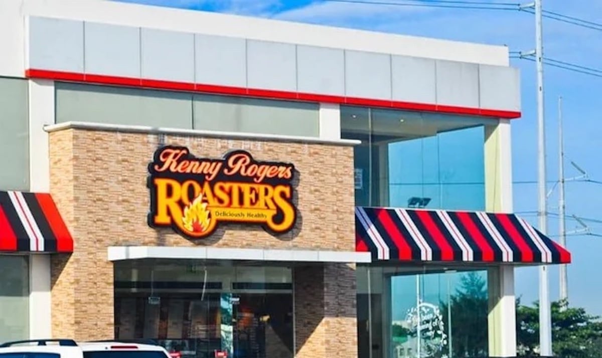 A Kenny Rogers Roasters, one of the classic old restaurant chains, with a large sign on a brick facade, features red, black, and white striped awnings and glass windows reflecting the sky and trees.