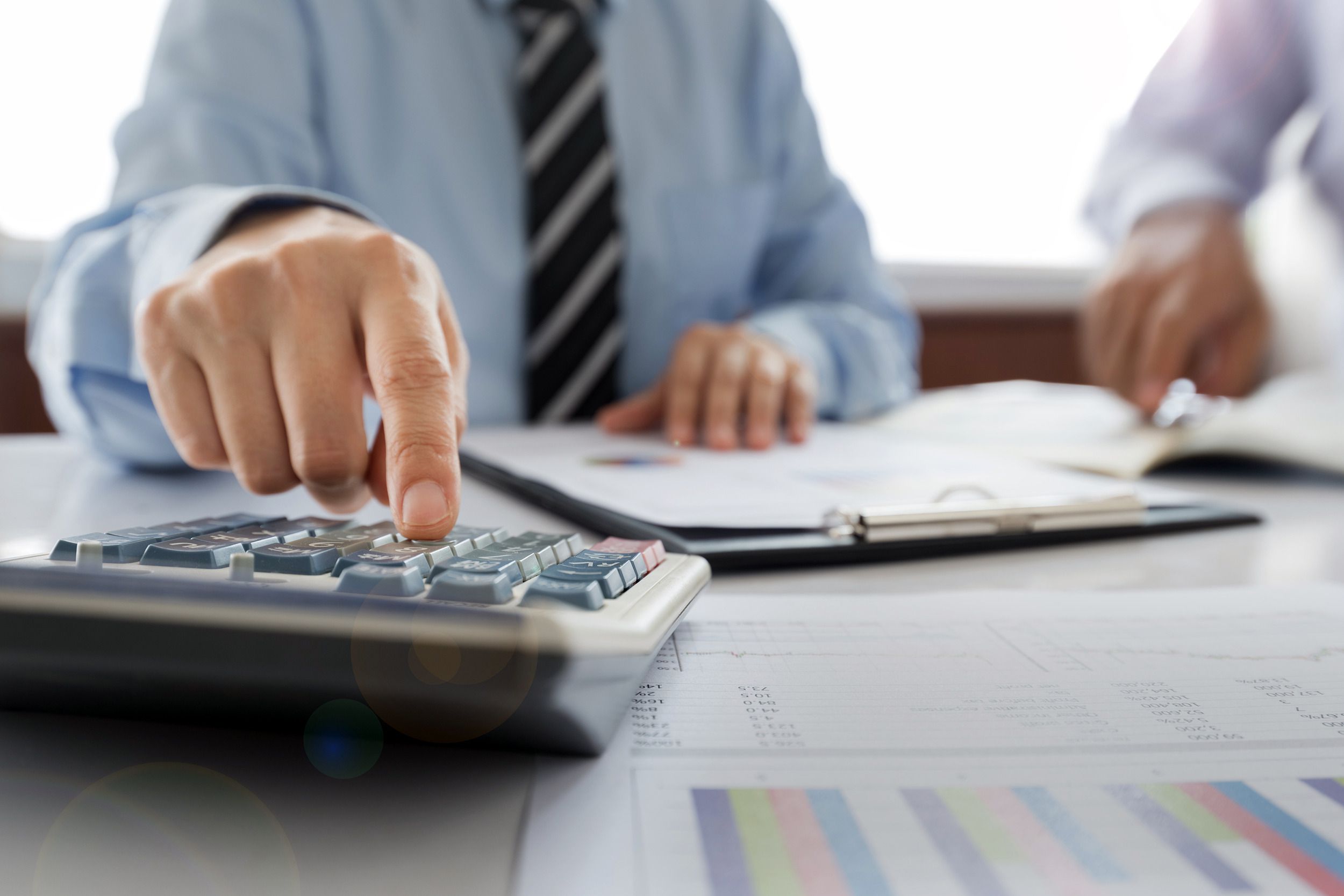 Male accountant with one hand using calculator, closeup, while other hand is on clipboard with papers, wearing blue shirt and striped tie, blurred background