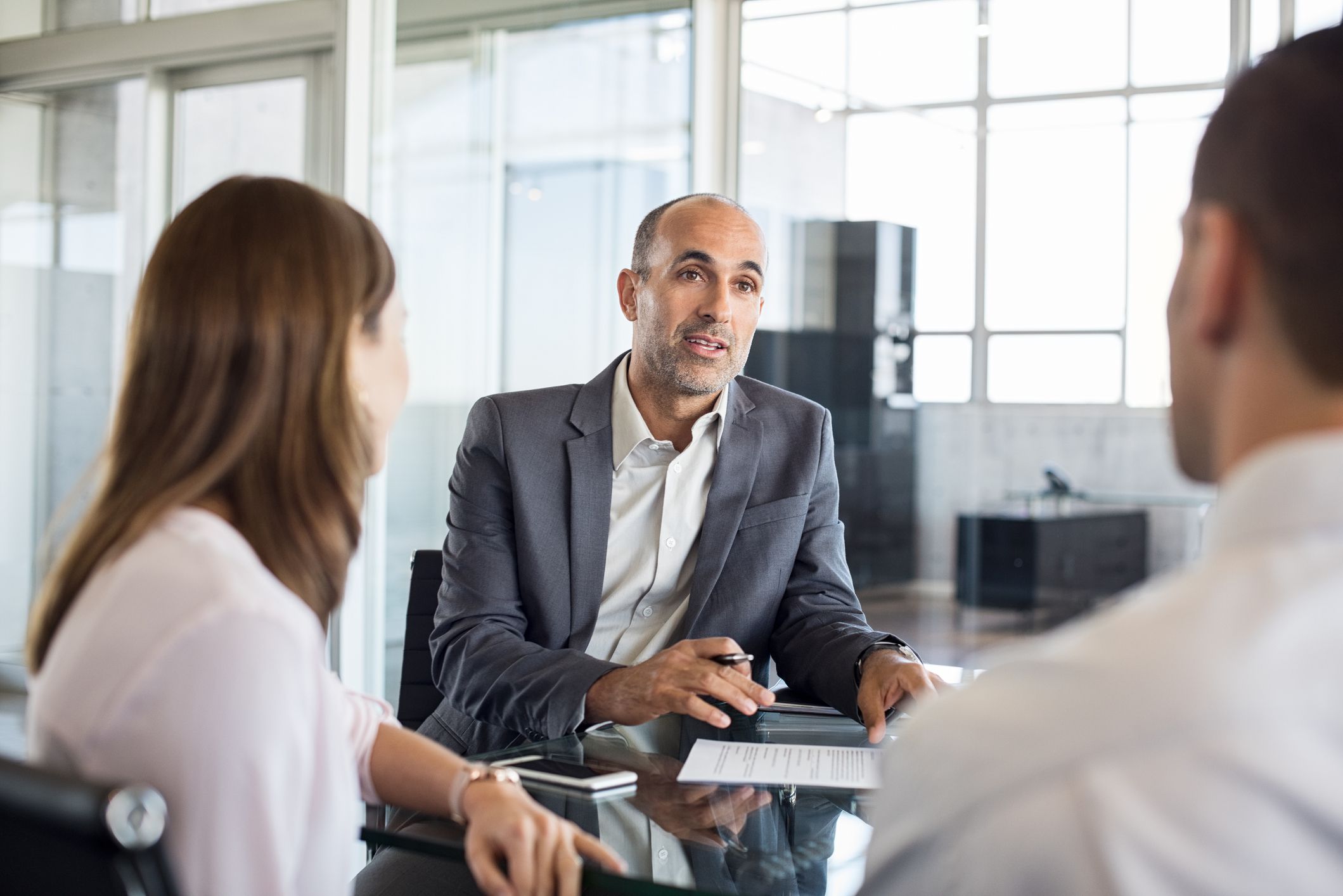 Male loan officer speaking to a couple, all are sitting at desk, office area in background