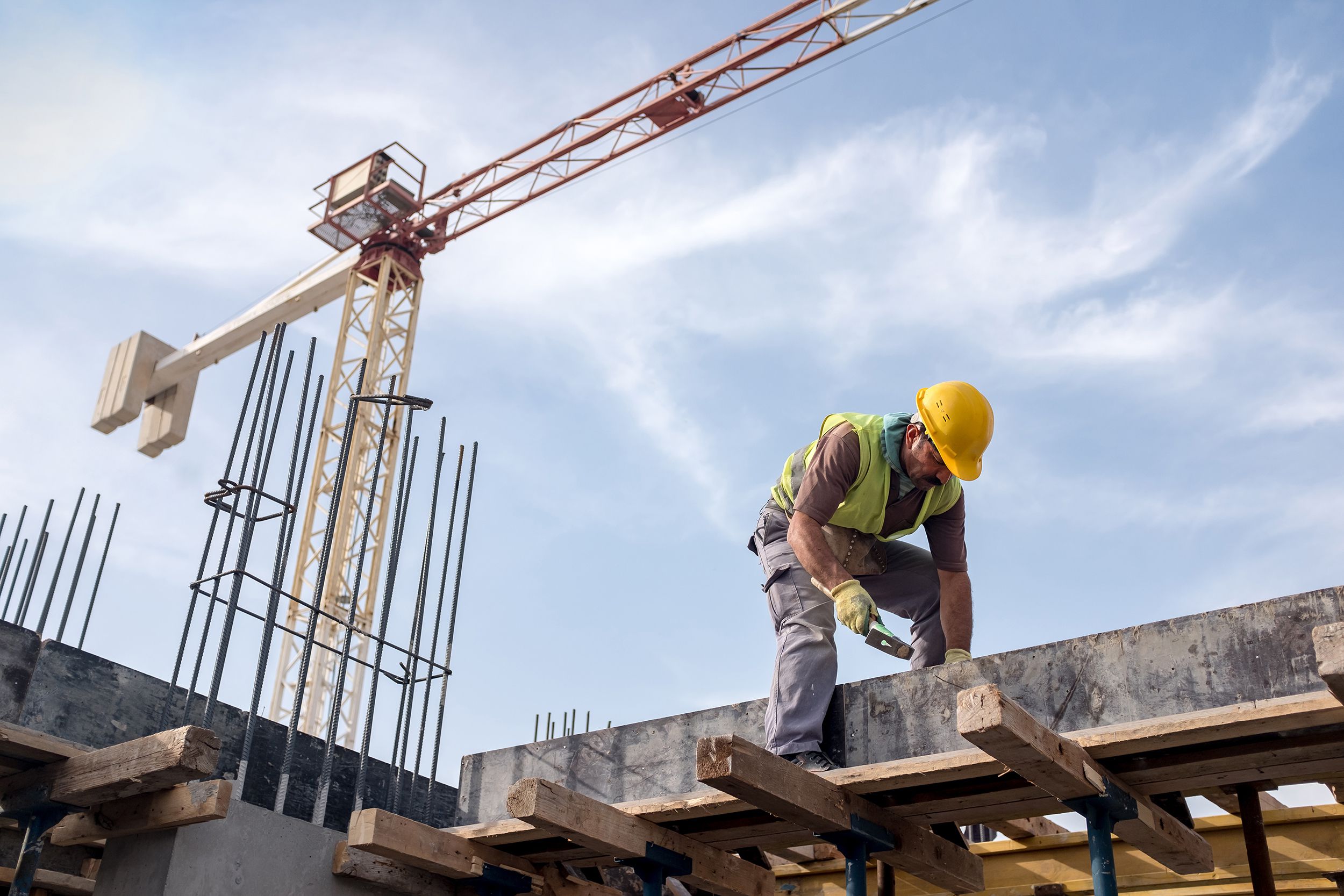 Male worker at construction site is fixing the form for the beam, he is standing on support as he is fixing from above, crane and sky is behind him