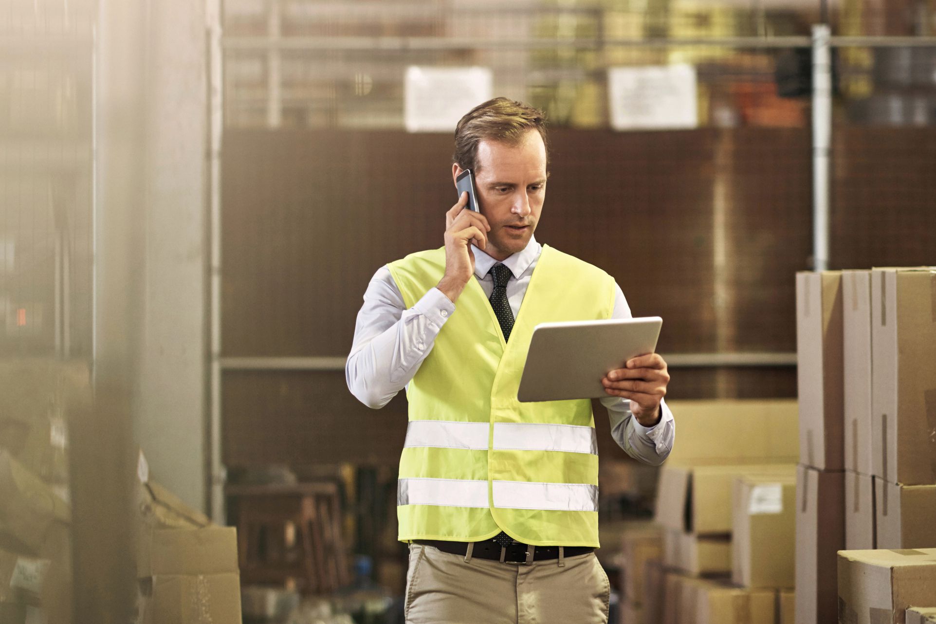 Man talking on mobile phone and using tablet in distribution warehouse, blurred background of boxes in warehouse