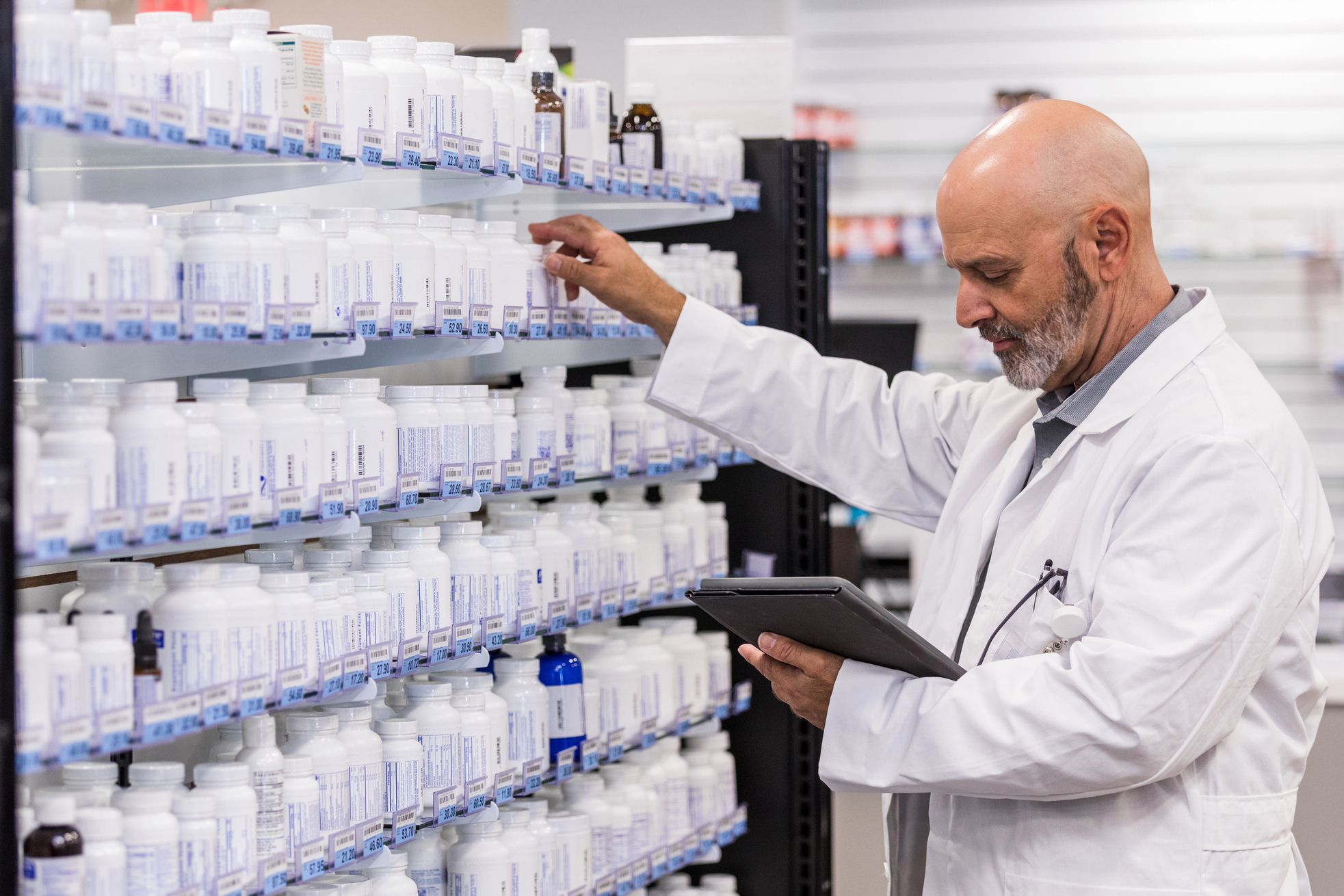Middle-aged male pharmacist checking inventory using a tablet, looking at tablet while touching bottles of medicine, blurred background of inside pharmacist medicine section