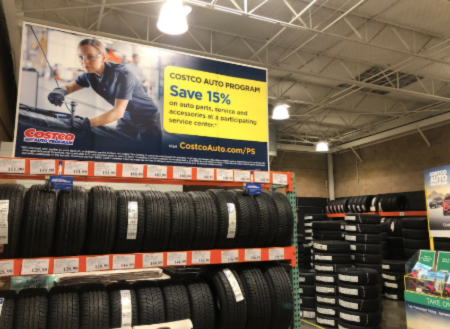 Rows of car tires are stacked on shelves in a warehouse store. A large sign above promotes the Costco Auto Program, offering 15% savings on auto parts, service, and accessories.