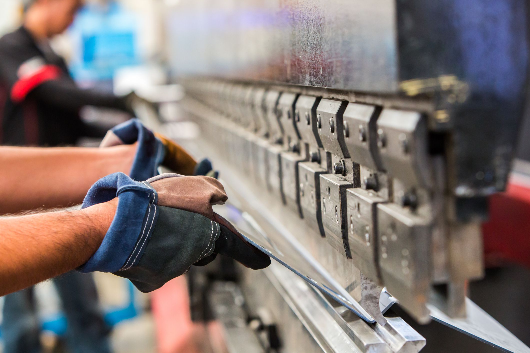 Person holding sheet metal while it is being bended by machine, blurred background of factory and two workers