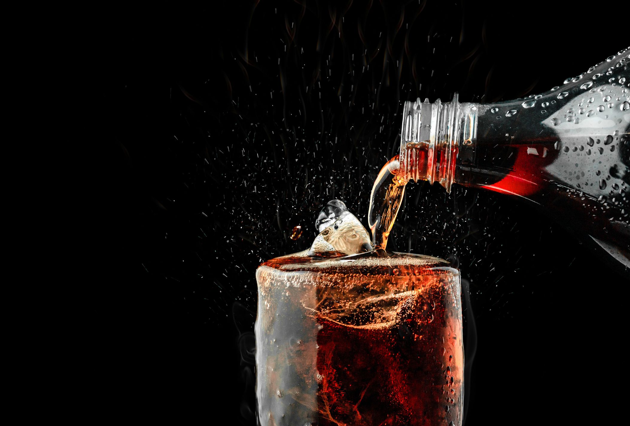 Side-view of dark soda being poured into a glass with ice, on right, clipped-view of soda bottle, against a black background