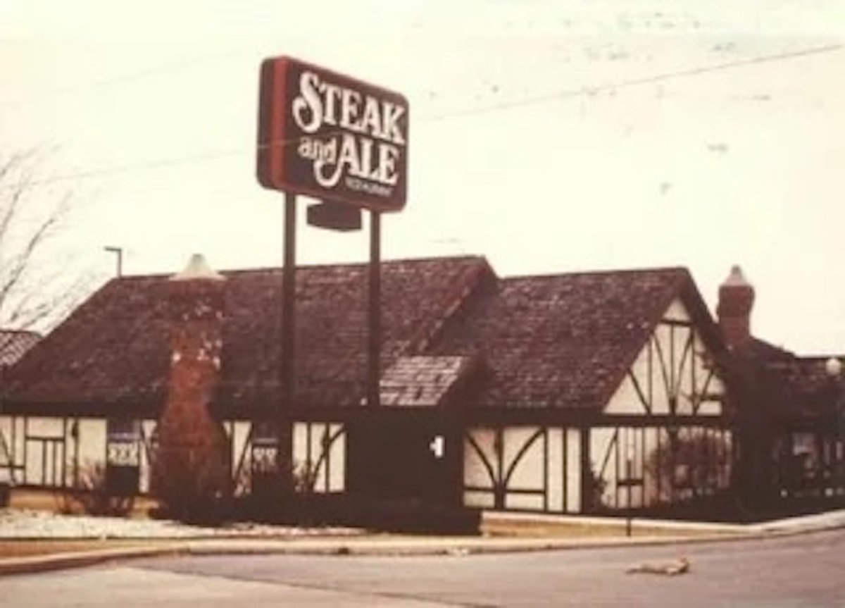 A vintage Steak and Ale from the era of old restaurant chains, with Tudor-style architecture, steep shingled roof, brick chimneys, and a large roadside sign. The scene is overcast and quiet.